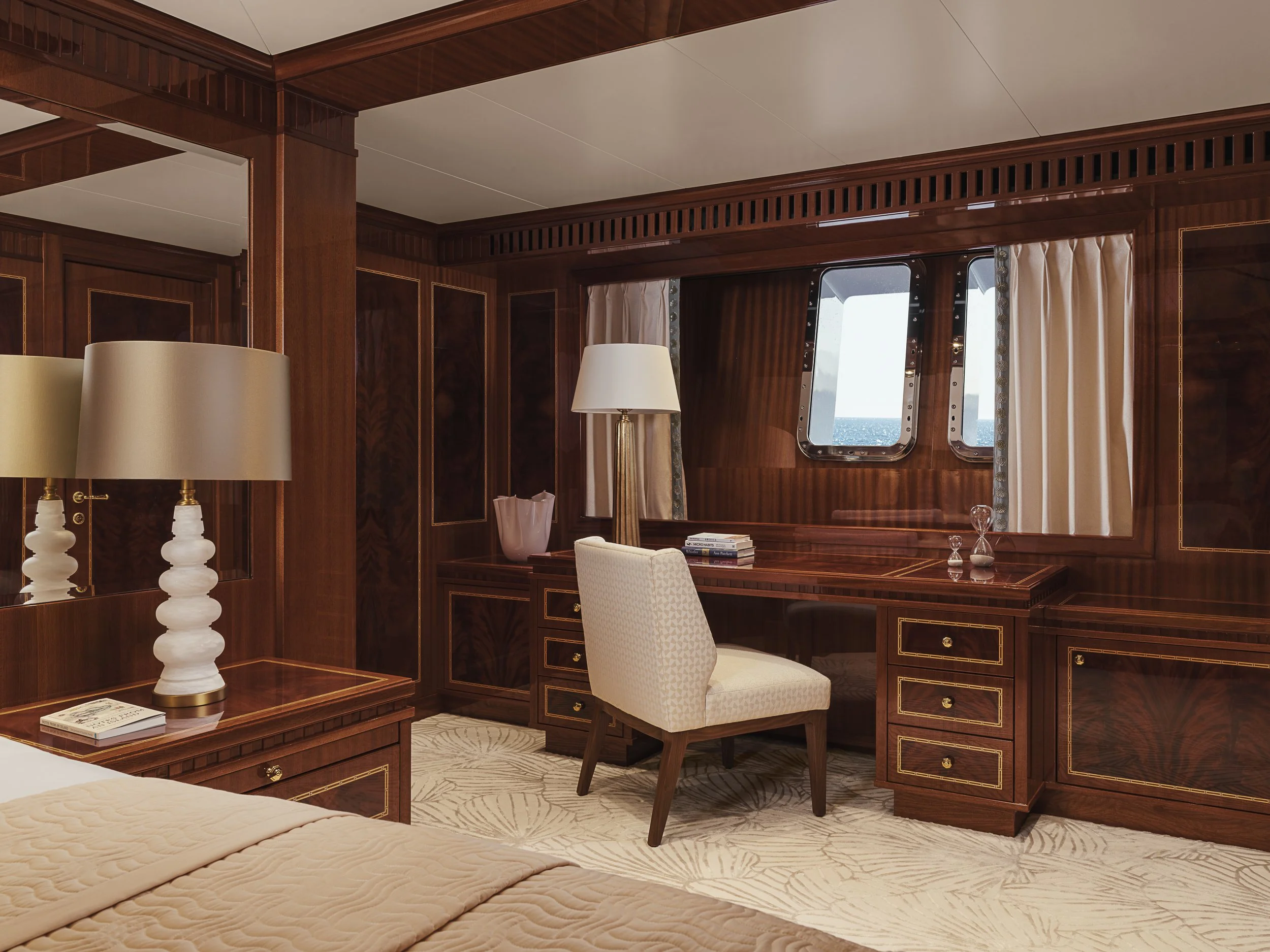 Interior view of a luxurious room with wooden walls and furniture, featuring a desk, a white upholstered chair, decorative lamps, and windows with cream-colored curtains showing a view of the ocean.