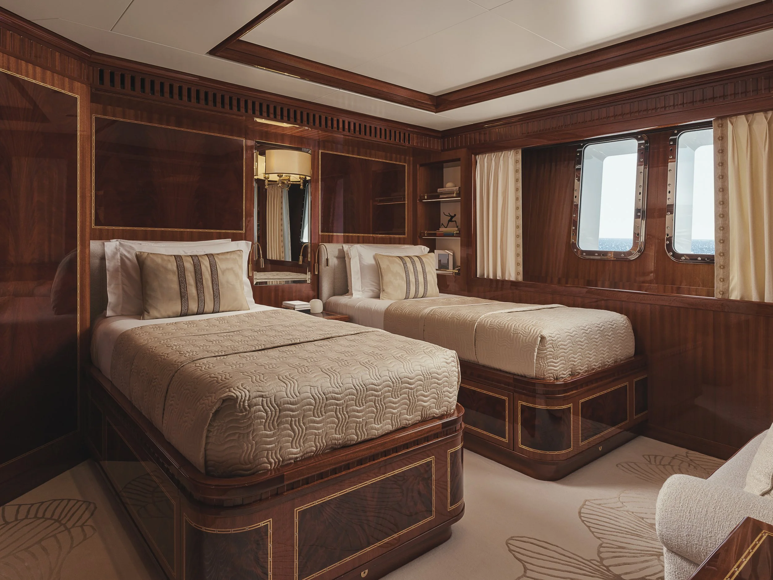 A luxurious bedroom with two twin beds made with cream blankets and beige pillows, featuring dark wood paneling and trim, with a mirror, books, a sculpture, and large windows with cream curtains overlooking the sea.