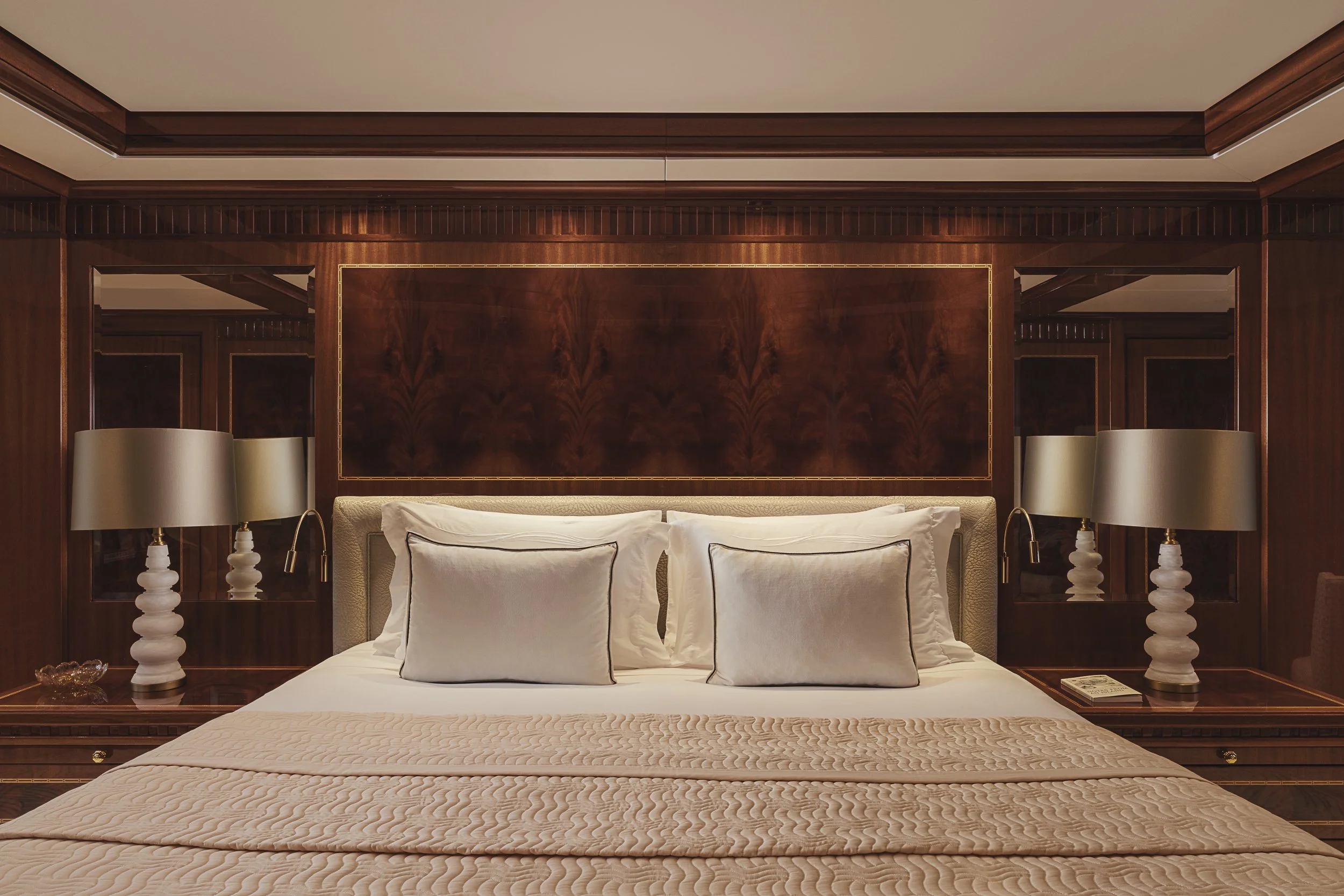A luxurious bedroom features a large bed with multiple white pillows and a beige quilted bedspread. The headboard, crafted from polished dark wood, extends across the width of the bed and is topped with a matching wooden panel decorated with gold tri