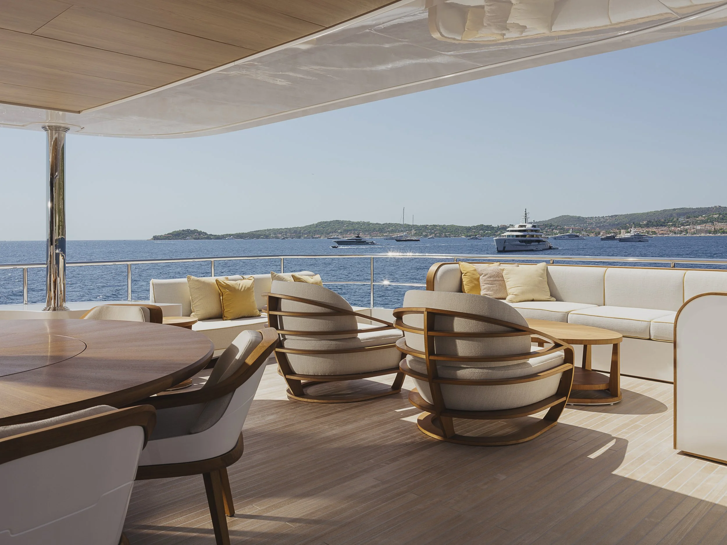 View from a yacht deck showing a seating area with white and beige cushioned chairs and wooden furniture, overlooking the ocean with several boats and an island in the background under a clear blue sky.