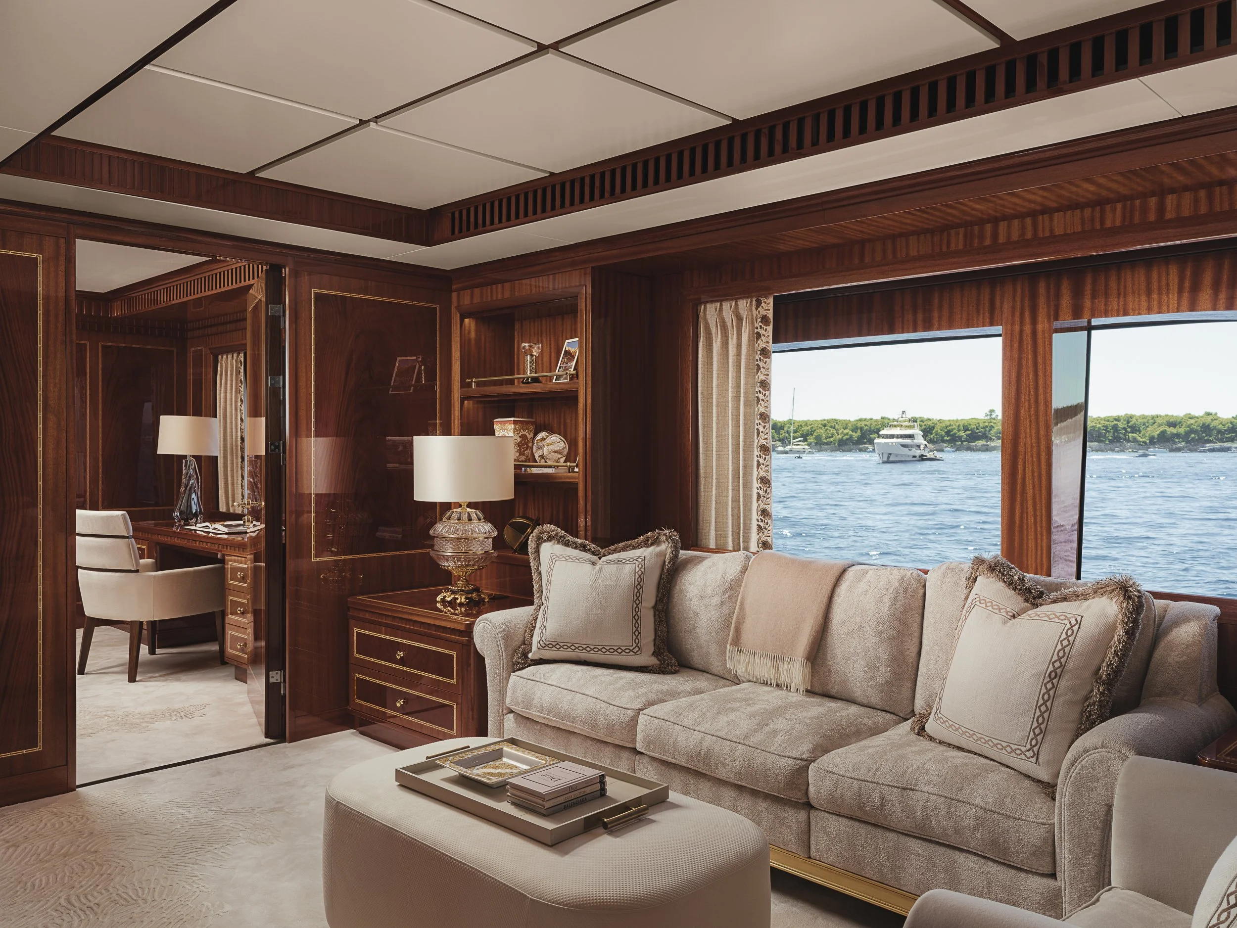 Interior view of a luxurious yacht or house with wood-paneled walls, large windows showing water and boats outside, and elegant furniture including a beige sofa with decorative pillows, a white ottoman, and a side table with books and a tray.