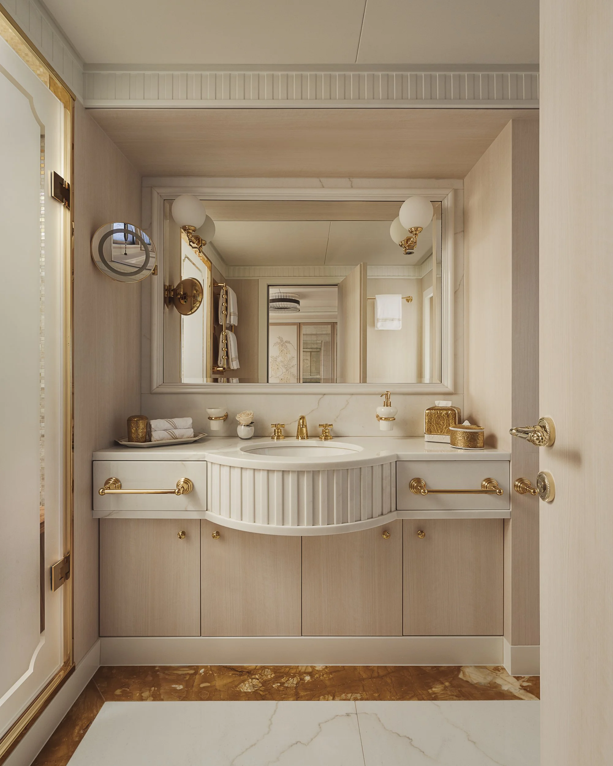 Elegant bathroom vanity with a large mirror, white countertop, gold fixtures, and decorative items including towels and small containers, with marble flooring.