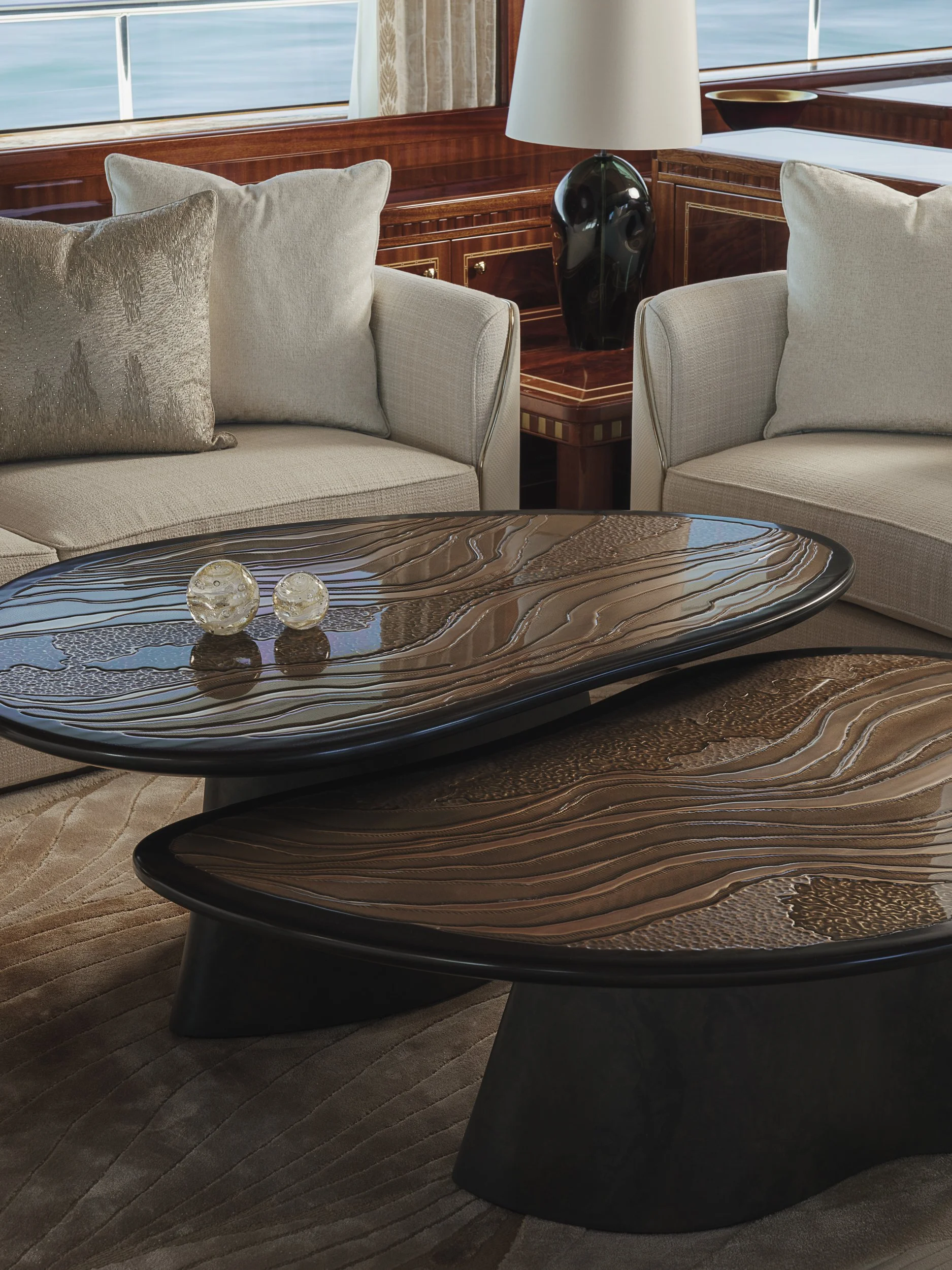 Living room with beige sofas, a wooden coffee table with a swirling pattern, a table lamp, and a window showing water outside.