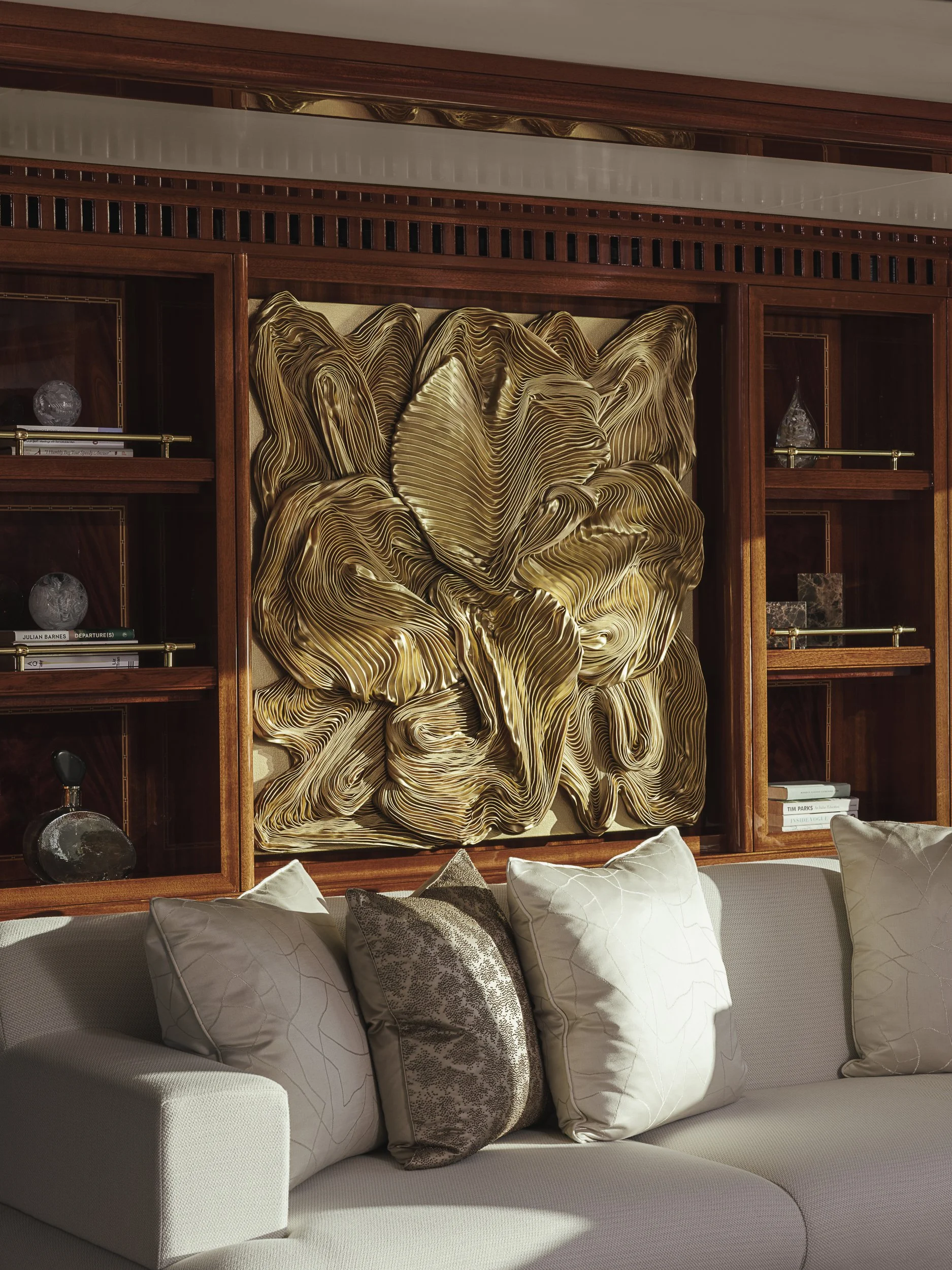 A beige sofa with decorative pillows in front of wooden bookshelves with decorative objects, including glass sculptures and books. A large abstract gold and beige artwork is mounted on the wall.