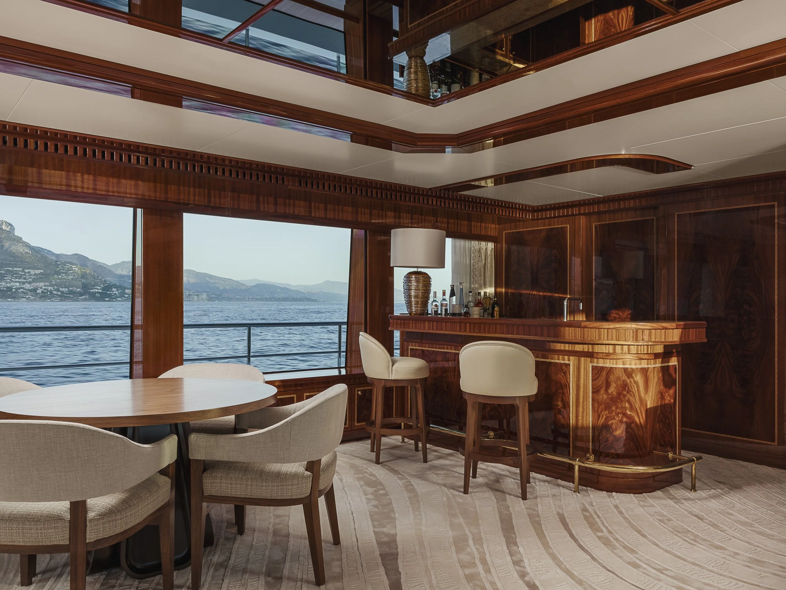 Interior of a luxurious boat or yacht with wood-paneled walls, a bar area with liquor bottles, a large round dining table with chairs, and a view of water and mountains outside.