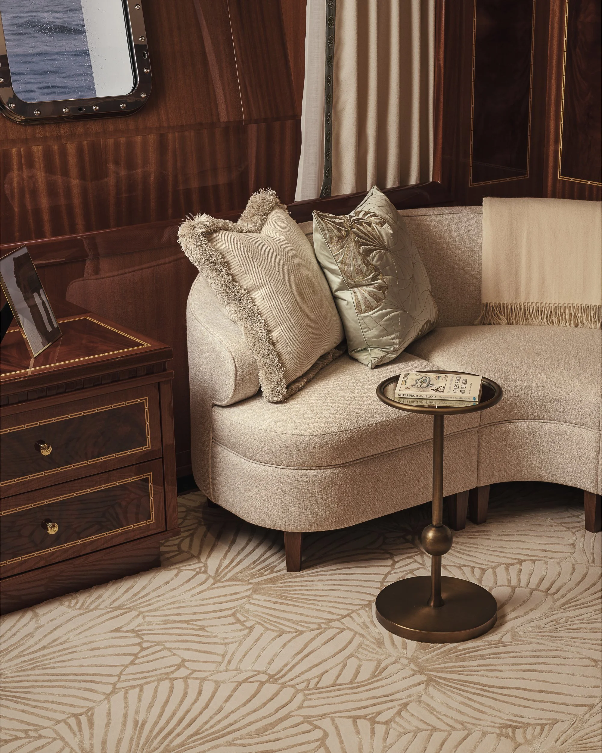 Luxurious interior with a beige curved sofa adorned with decorative pillows, a round side table with a book on it, a patterned rug, wooden panel walls, and a window with curtains.