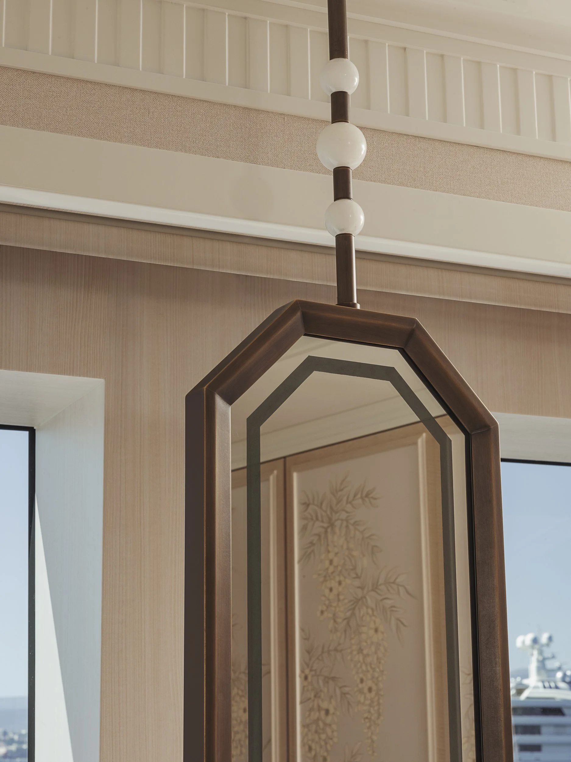 Close-up of a modern hanging light fixture with three round white globes and a wooden brown frame, mounted on a ceiling near a wall with a framed decorative art piece and window.