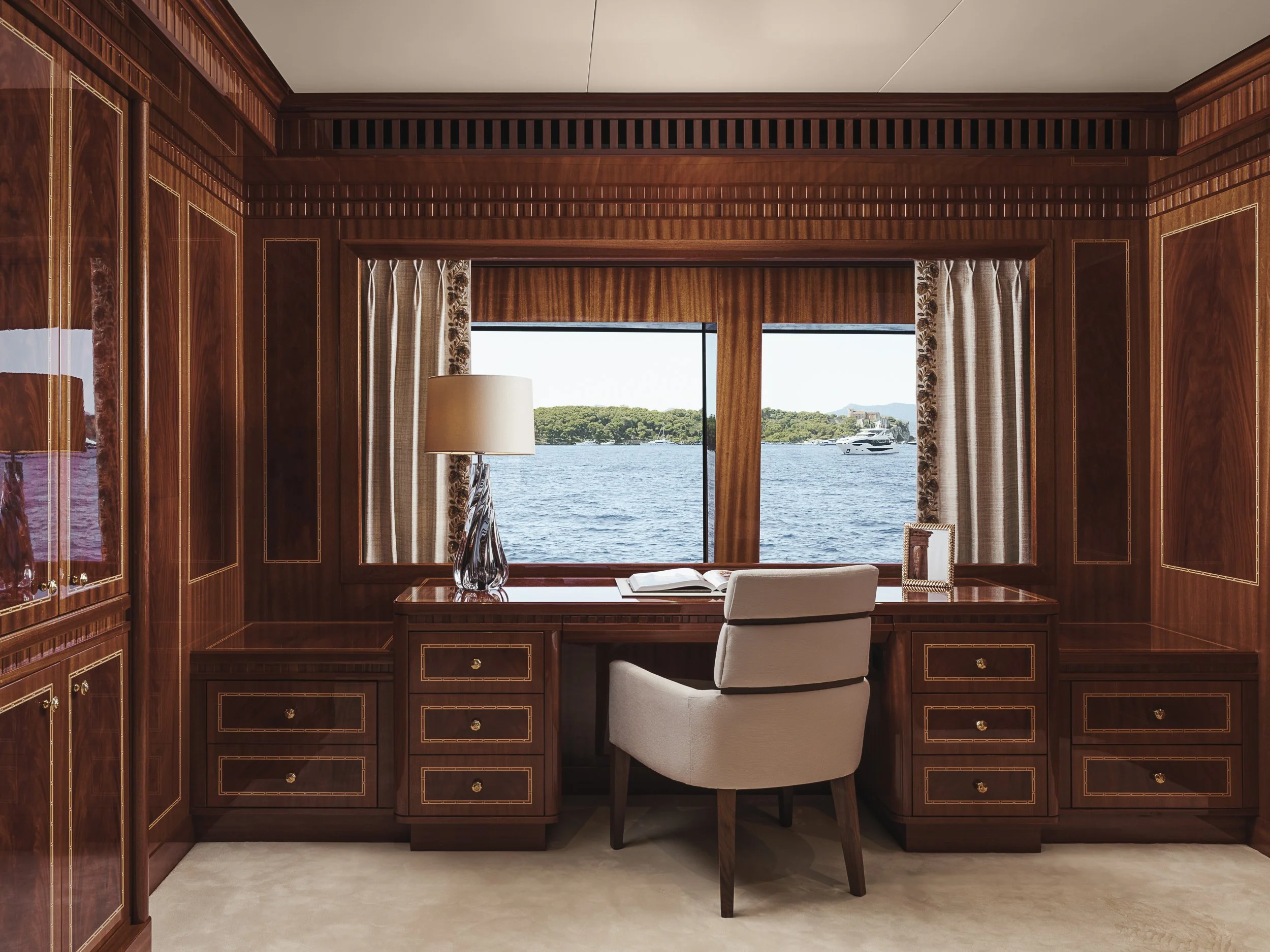 A room with wood-paneled walls and a view of water and greenery through a large window. There is a desk with a white chair, a book, a lamp, and curtains with floral patterns.