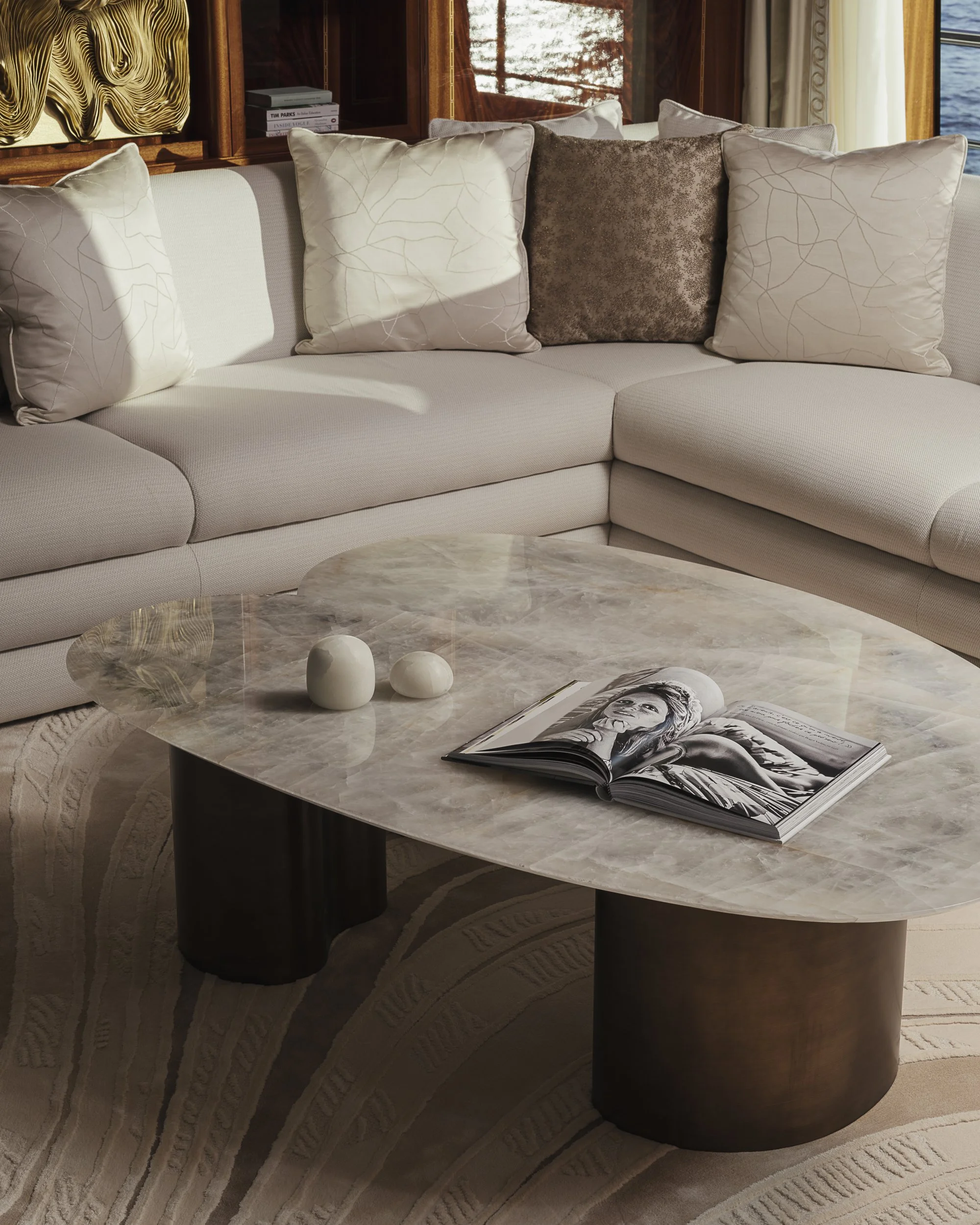 Living room with a light-colored sofa, five decorative pillows, a marble coffee table with a magazine, and a large window with a view of water outside.