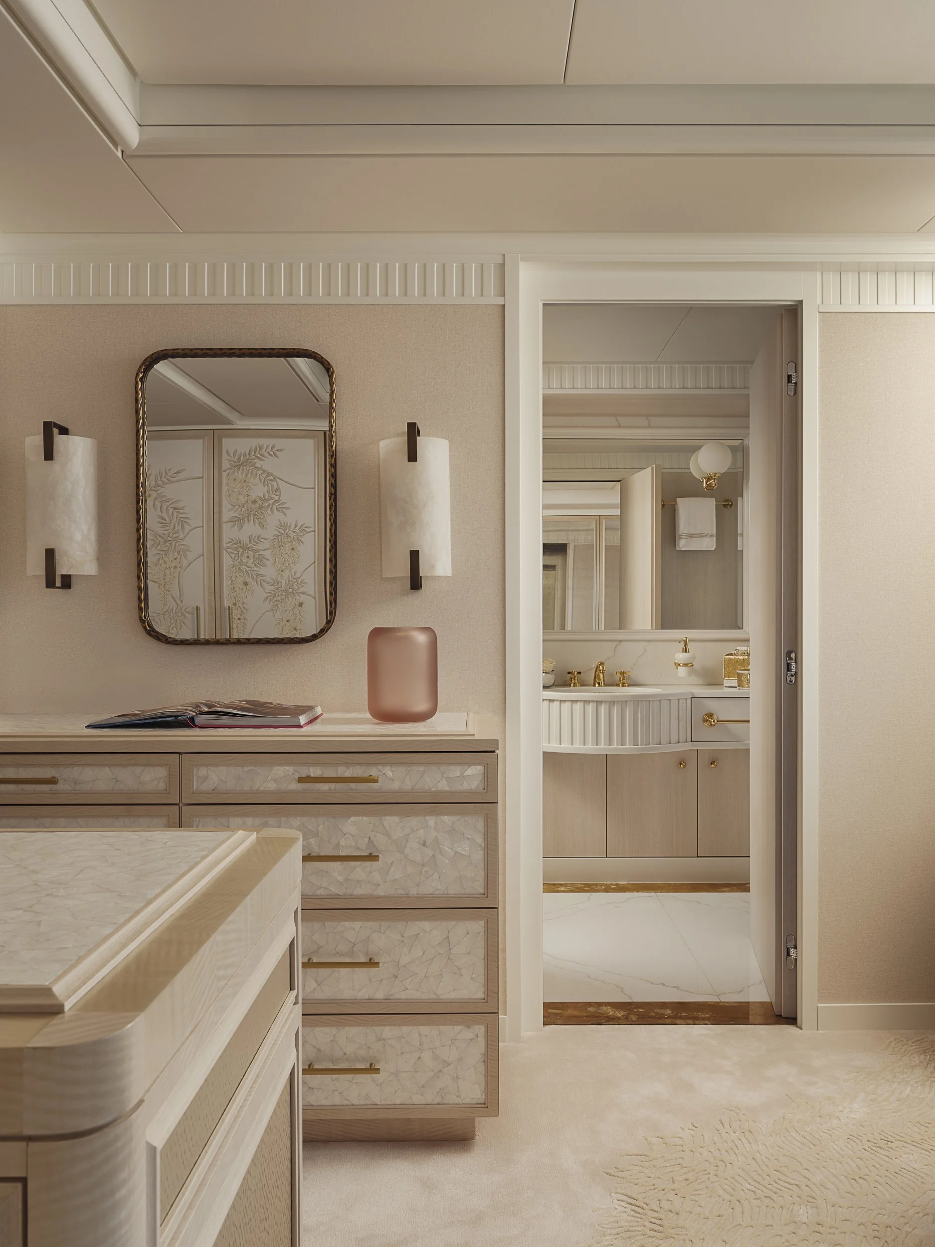 A hotel room with light-colored furniture, a dresser with gold handles, a mirror, wall-mounted lights, and a view of the bathroom through an open door. The bathroom has a marble countertop with gold fixtures and a mirror. The room has beige walls, a 