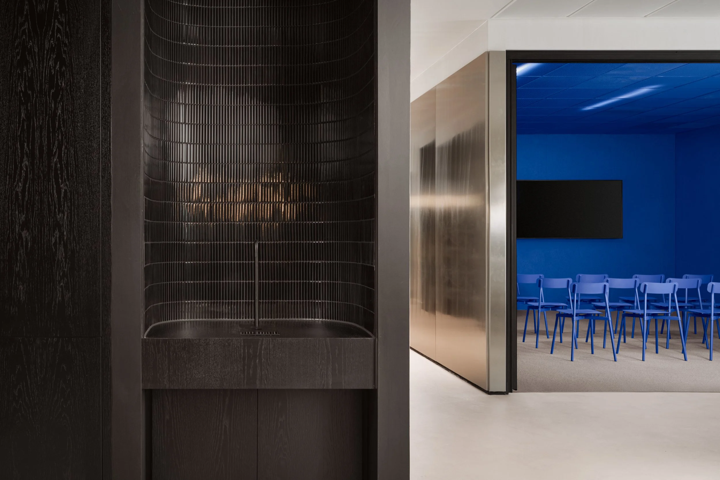 Modern interior space with a black, curved, wire mesh seating area on the left and a blue-walled conference room with multiple blue chairs and a large black screen on the right.