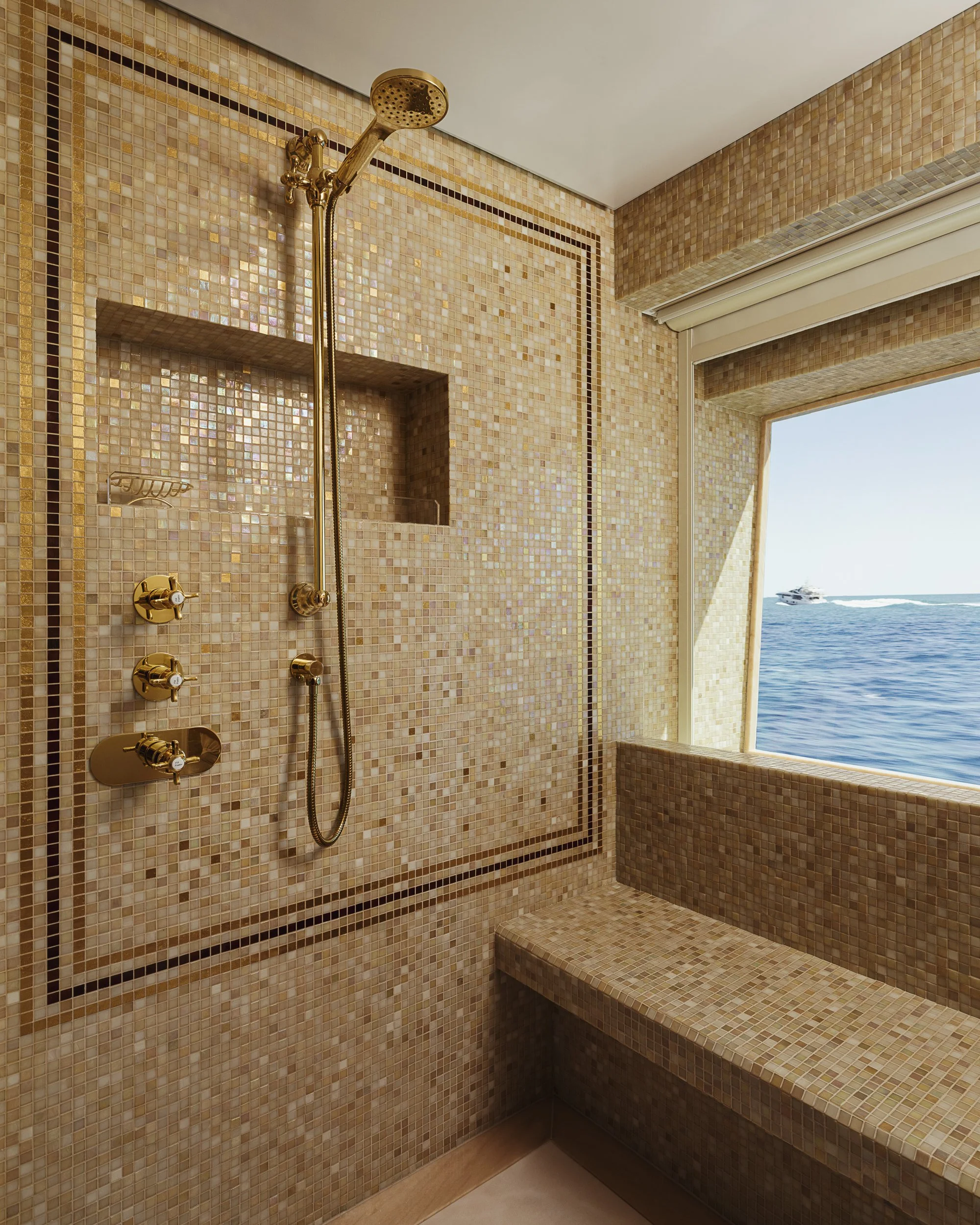 A luxurious shower area with gold fixtures, mosaic tile walls in shades of beige, pink, and gold, a built-in shelf, and a large window offering a view of the sea and a yacht outside.