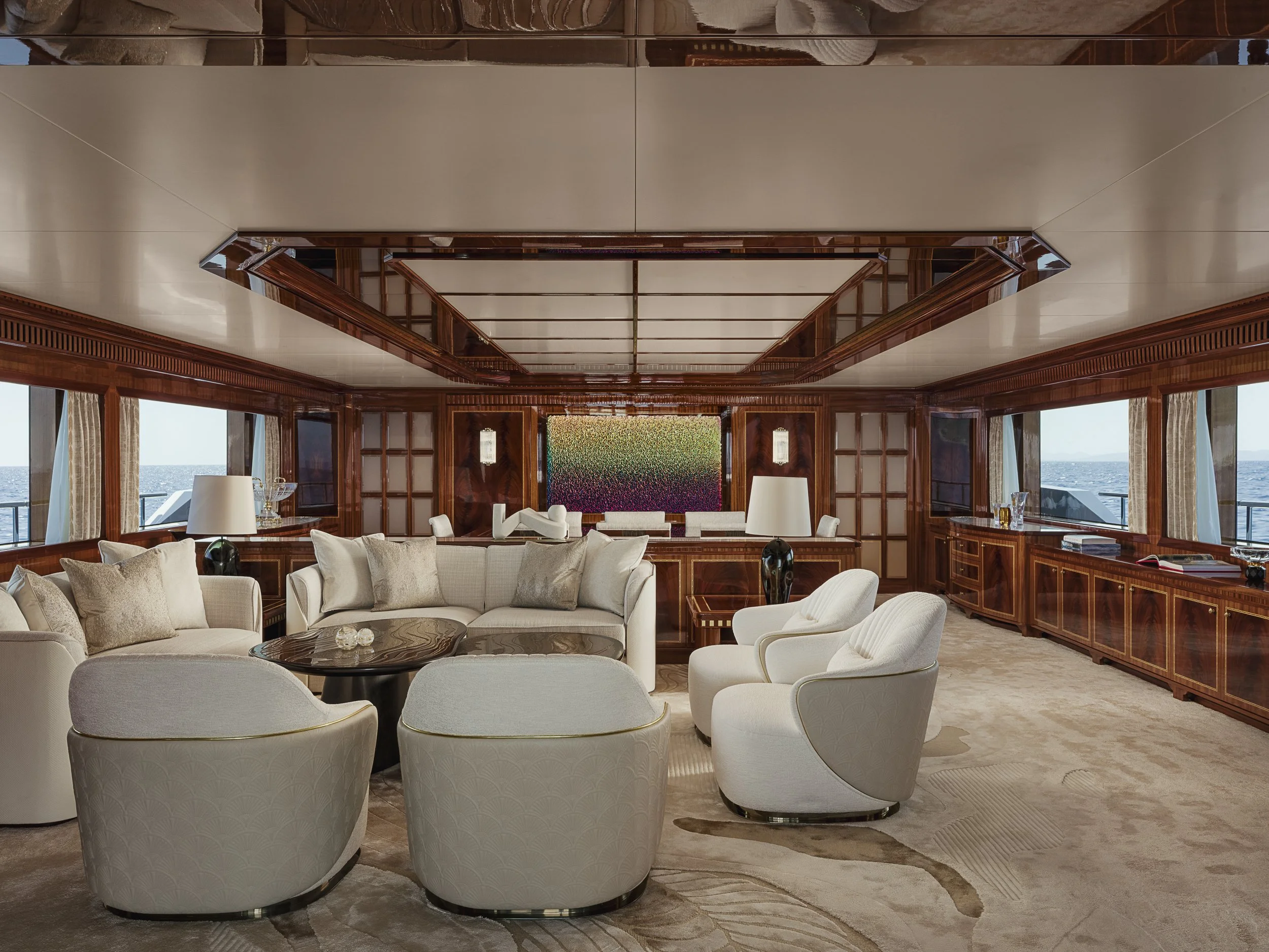 A luxurious living room with cream-colored seating arranged around a dark wooden coffee table. The room has large windows showing an ocean view, and the walls are decorated with dark wood paneling and ornate trim. There are white lamps on side tables