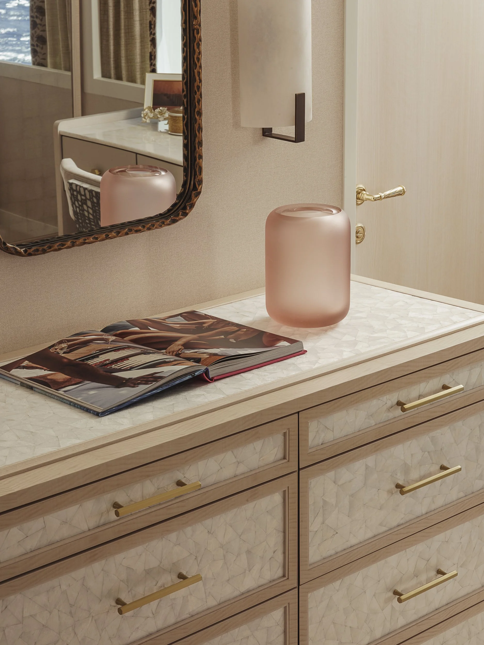 A marble-top wooden dresser with a gold drawer handle, a pink frosted glass lamp, an open magazine, a mirror with a leopard print frame, a small framed picture, and a decorative vase on top of it.