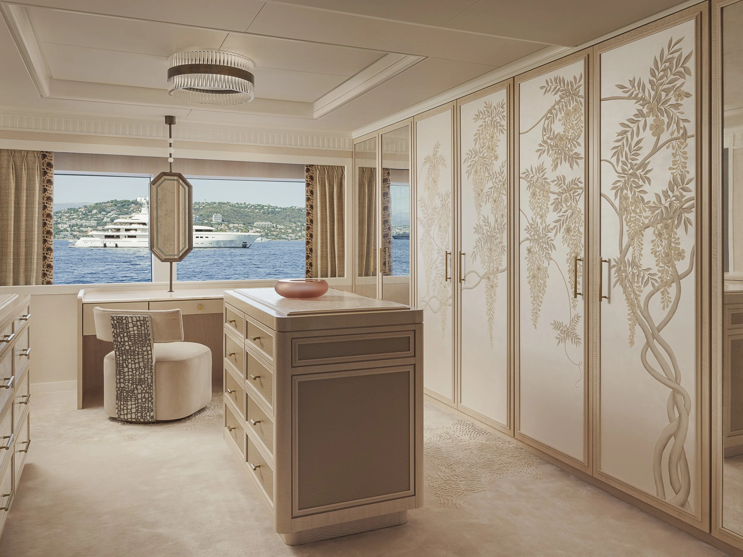 Luxury room with beige and cream furniture, a view of water and a yacht through the window, ornate wallpaper with a floral pattern, and elegant ceiling lighting.