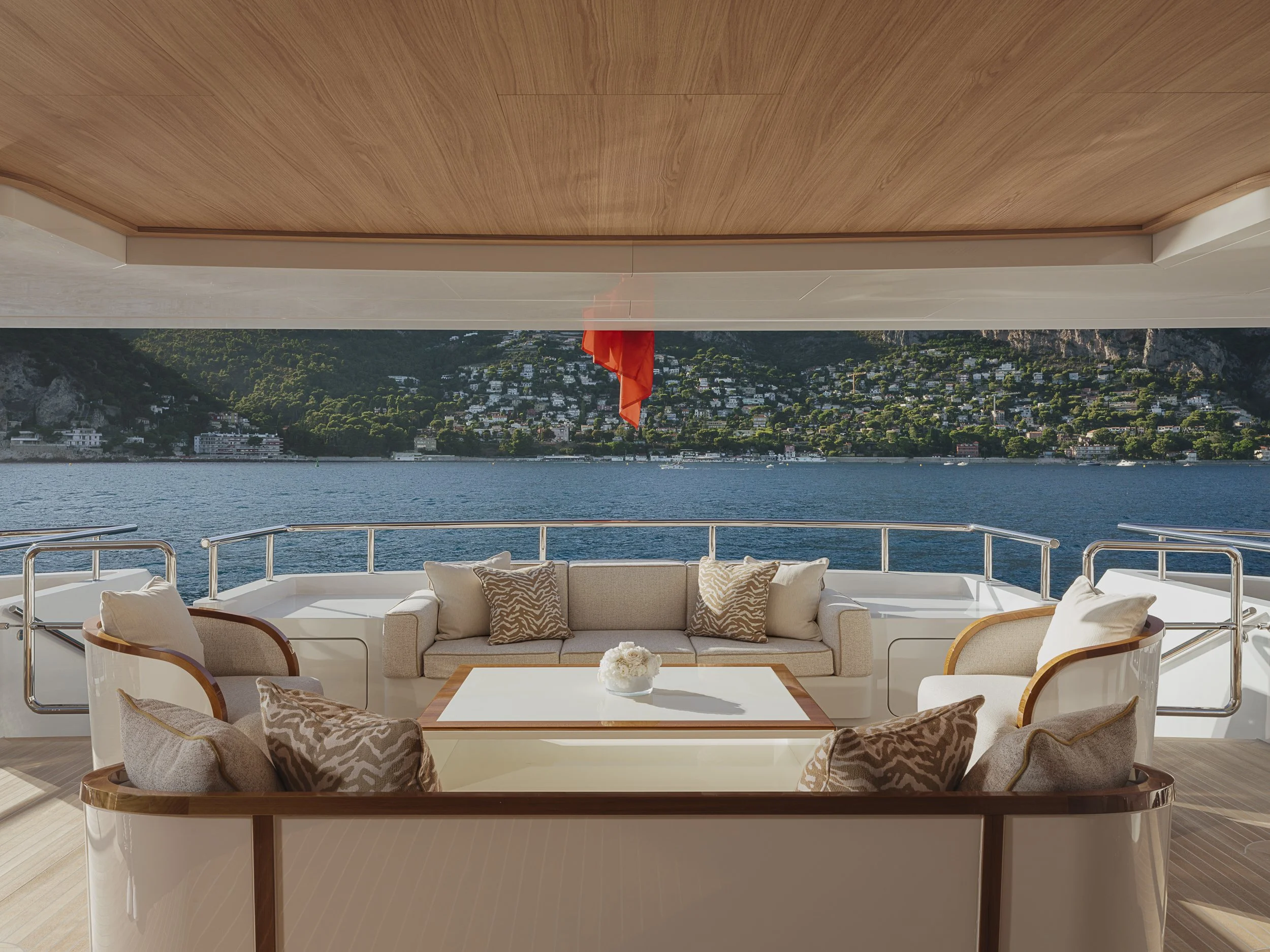 A luxurious yacht deck with a seating area, views of a waterfront and lush green hills, under a wooden ceiling, with some decorative pillows and a flower on the table.