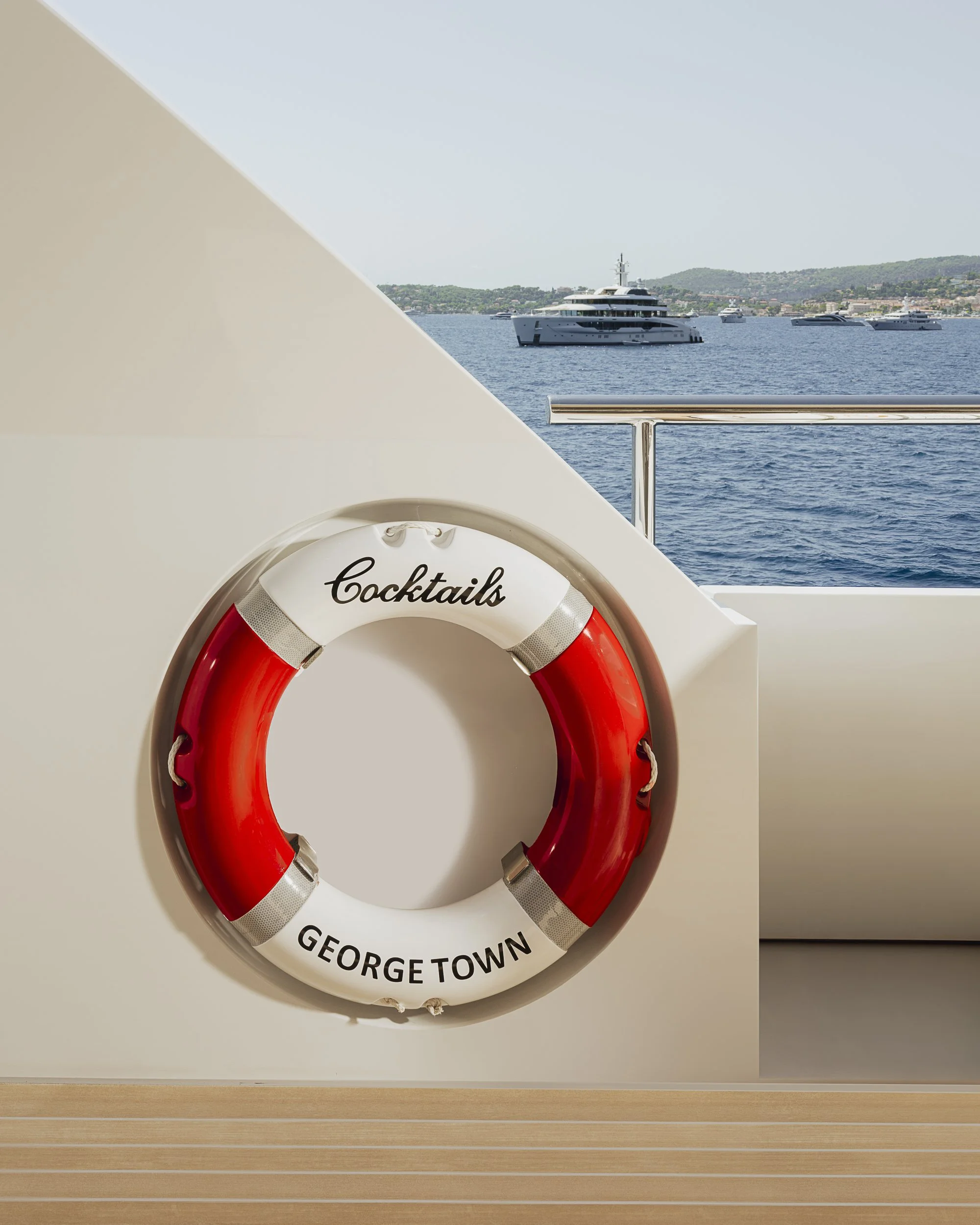 Life preserver ring labeled 'George Town' and 'Cocktails' hanging on a boat rail, with boats on the water and a coastline in the background.