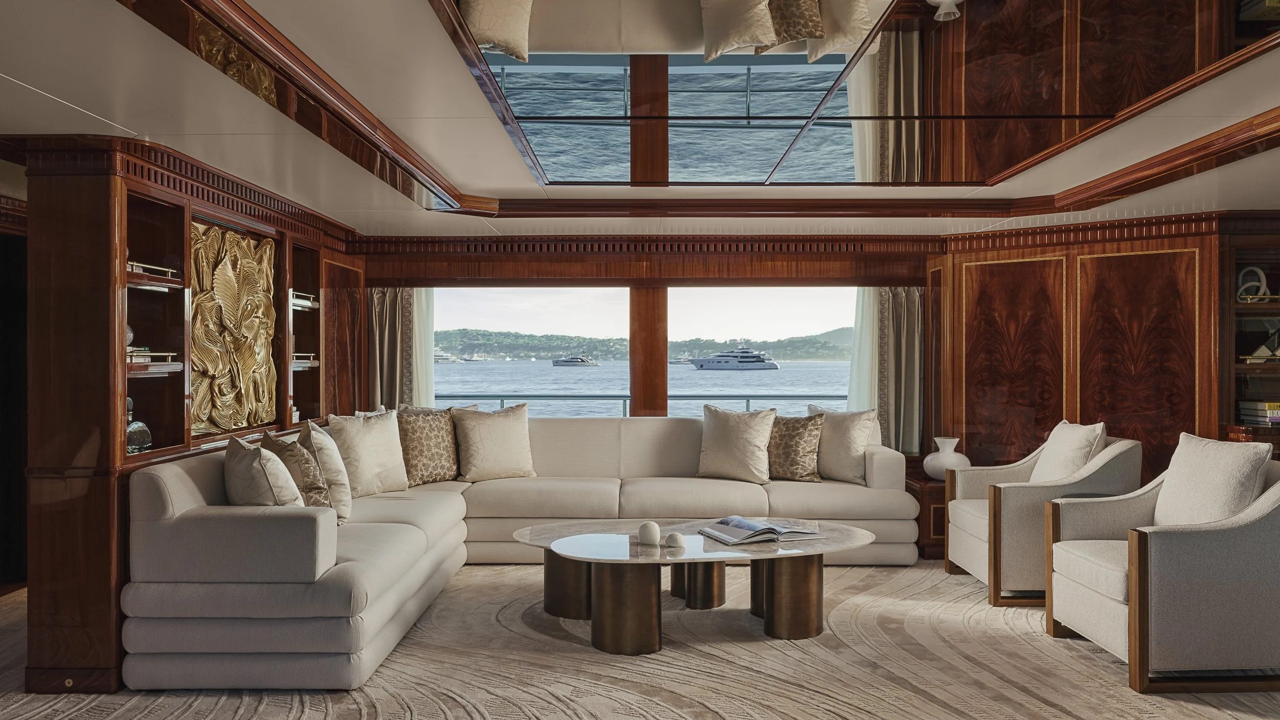 Living room with large window view of water and boats, white sectional sofa with beige and patterned pillows, round marble-top coffee table with open magazine and decorative objects, wooden built-in shelves and cabinetry, cream armchair, curtains on 