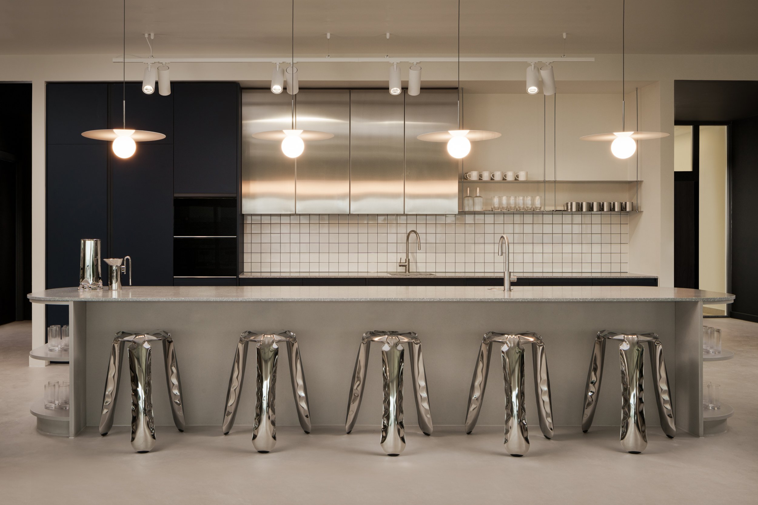 Modern kitchen with a large island, metallic bar stools, pendant lighting, black cabinets, white tiled backsplash, and minimal decor.