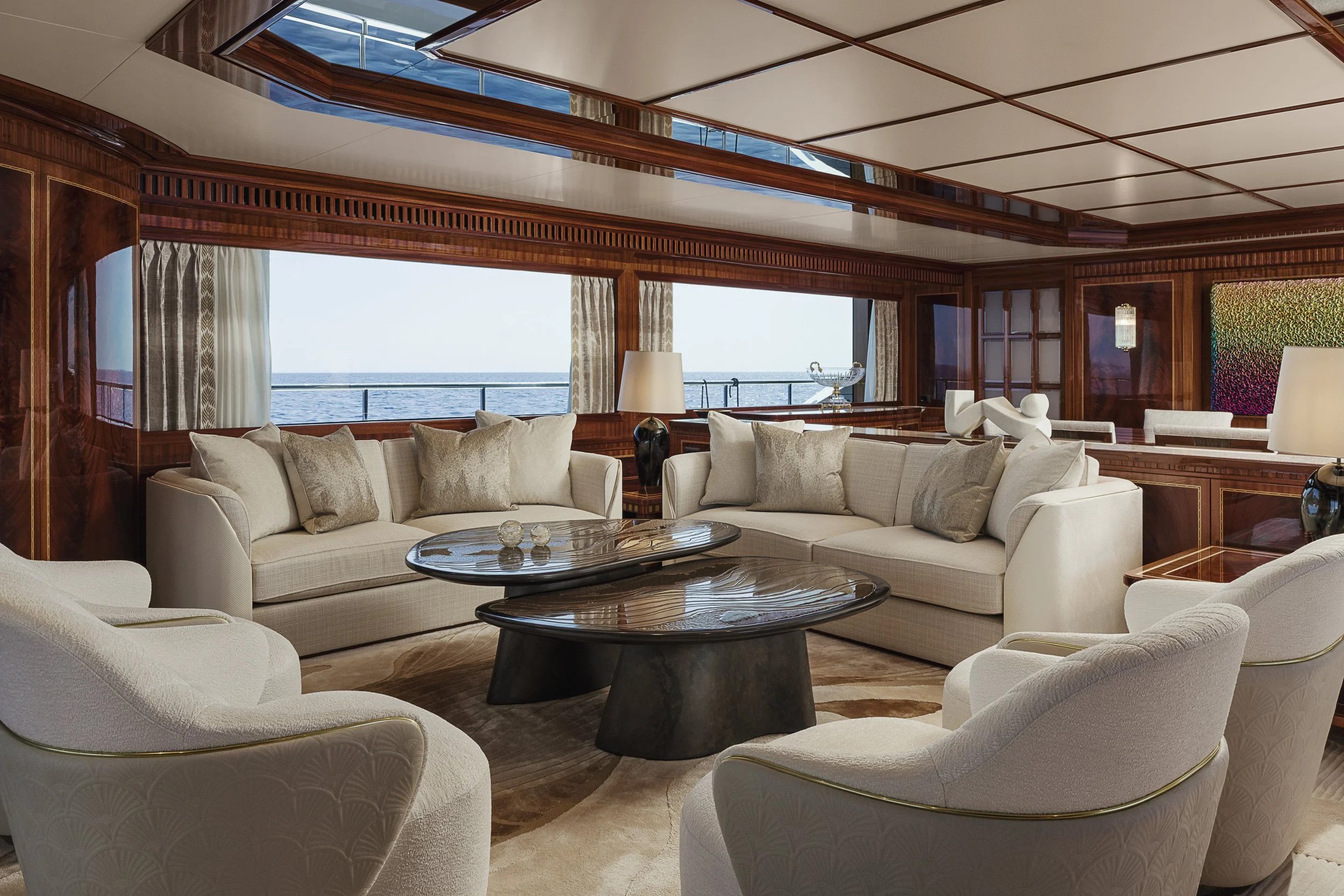 Luxury living room with cream-colored sofas, dark wood tables, large windows showing ocean view, and wooden ceiling panels.