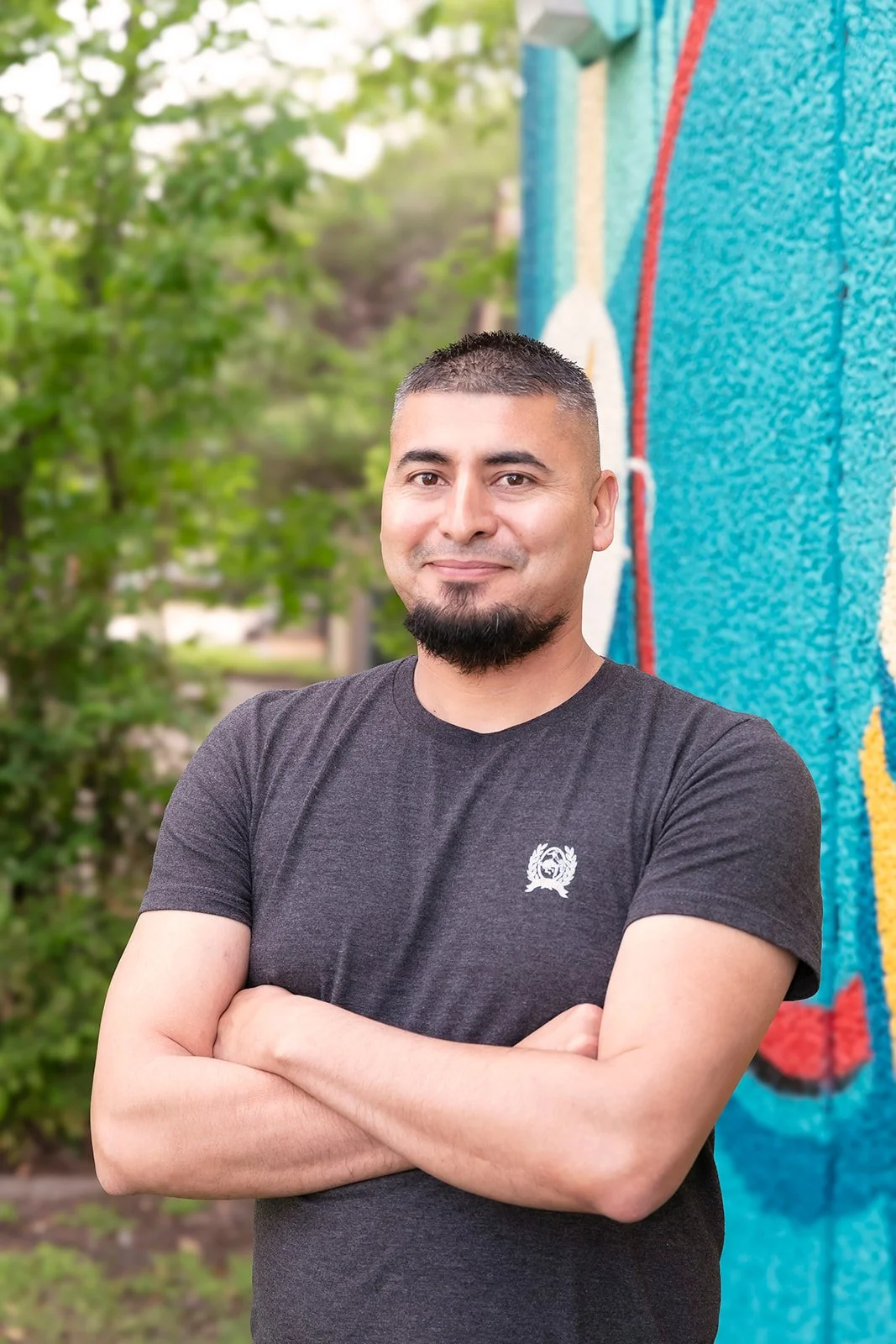 Close-up portrait of a young man with short black hair, a goatee, and wearing a black shirt, standing in front of a mural and trees..