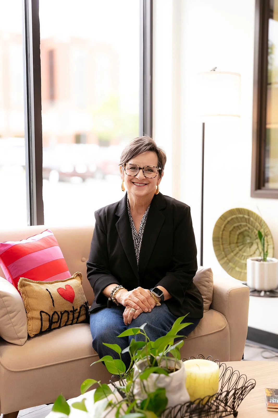 A woman with short gray hair, glasses, and a black blazer sitting on a beige couch near large windows, smiling, surrounded by colorful pillows and houseplants, in a bright, modern room.