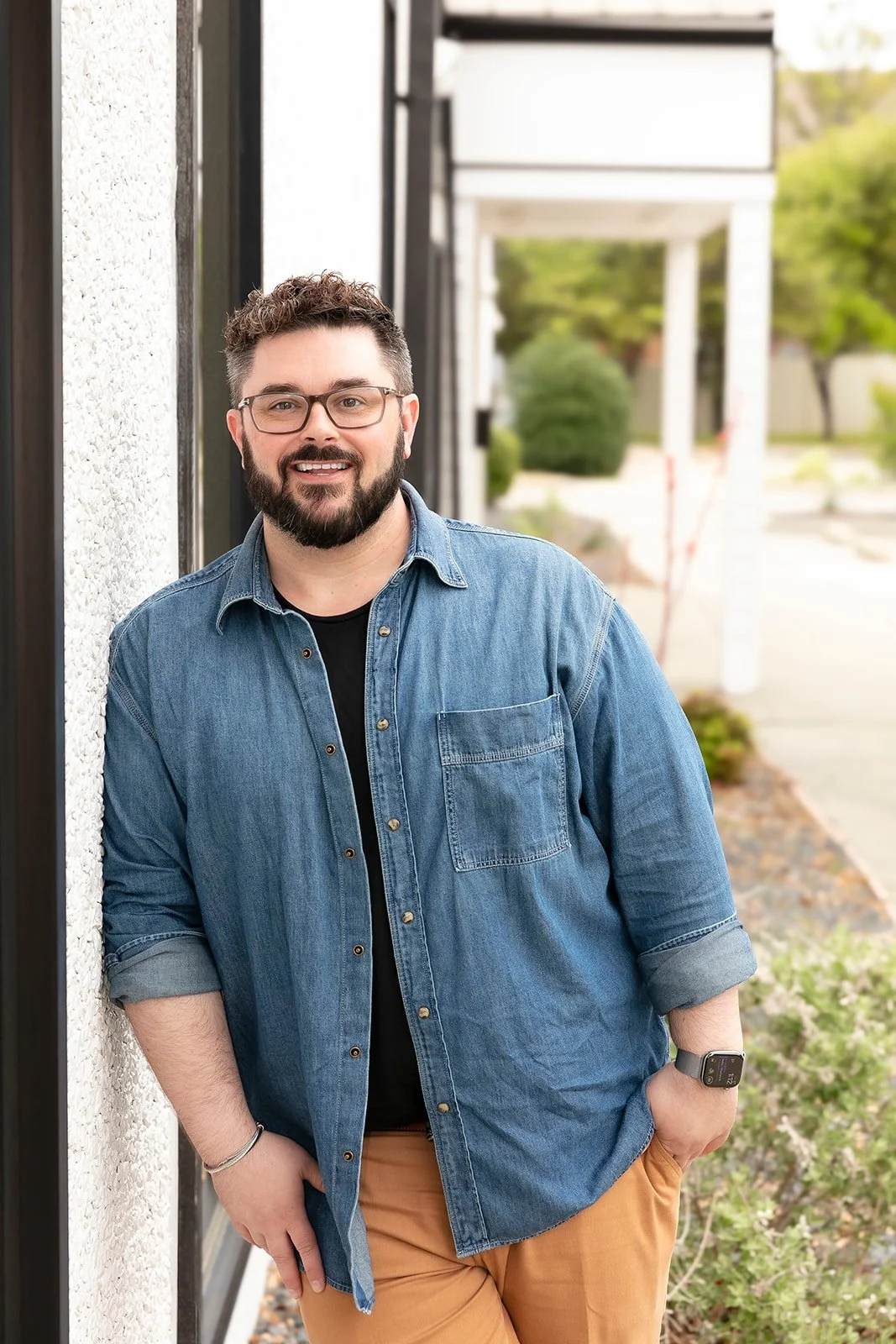 Hutch Ice smiling and leaning on an outside wall in a denim shirt. 