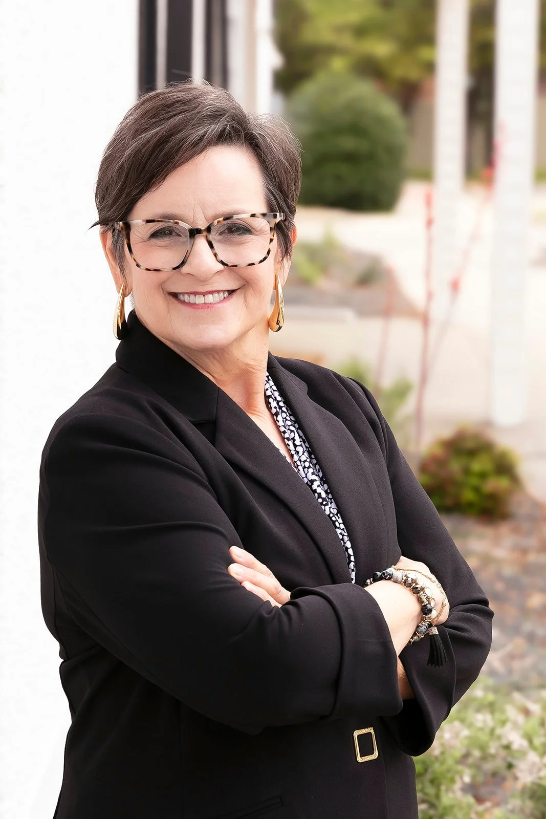 A smiling woman with short dark hair and glasses, wearing a black Leans on a wall. 