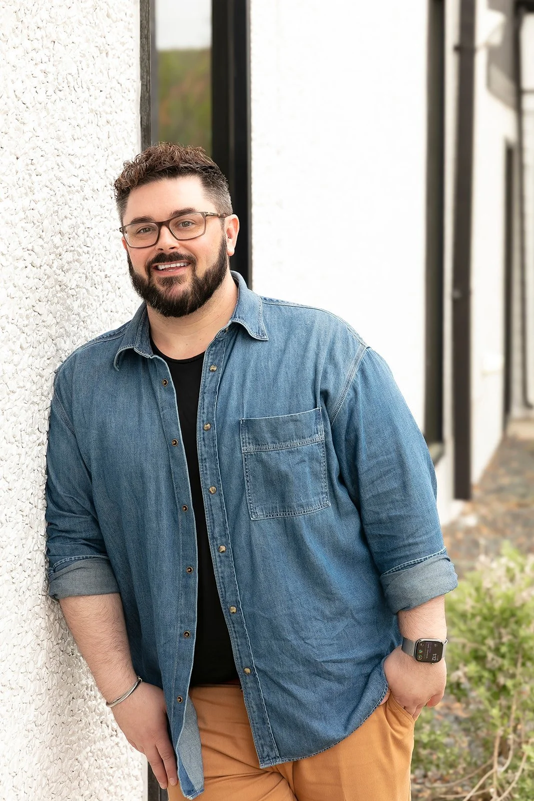 A smiling man with glasses, a beard, and short curly hair standing outdoors next to a textured white wall, wearing a denim shirt, black t-shirt, tan pants, and a smartwatch.