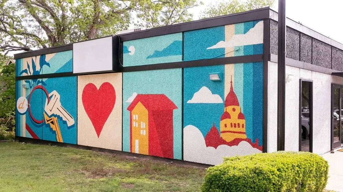 Colorful mural on a building wall featuring abstract designs, a heart, a house, a church, and clouds in a sunny sky.