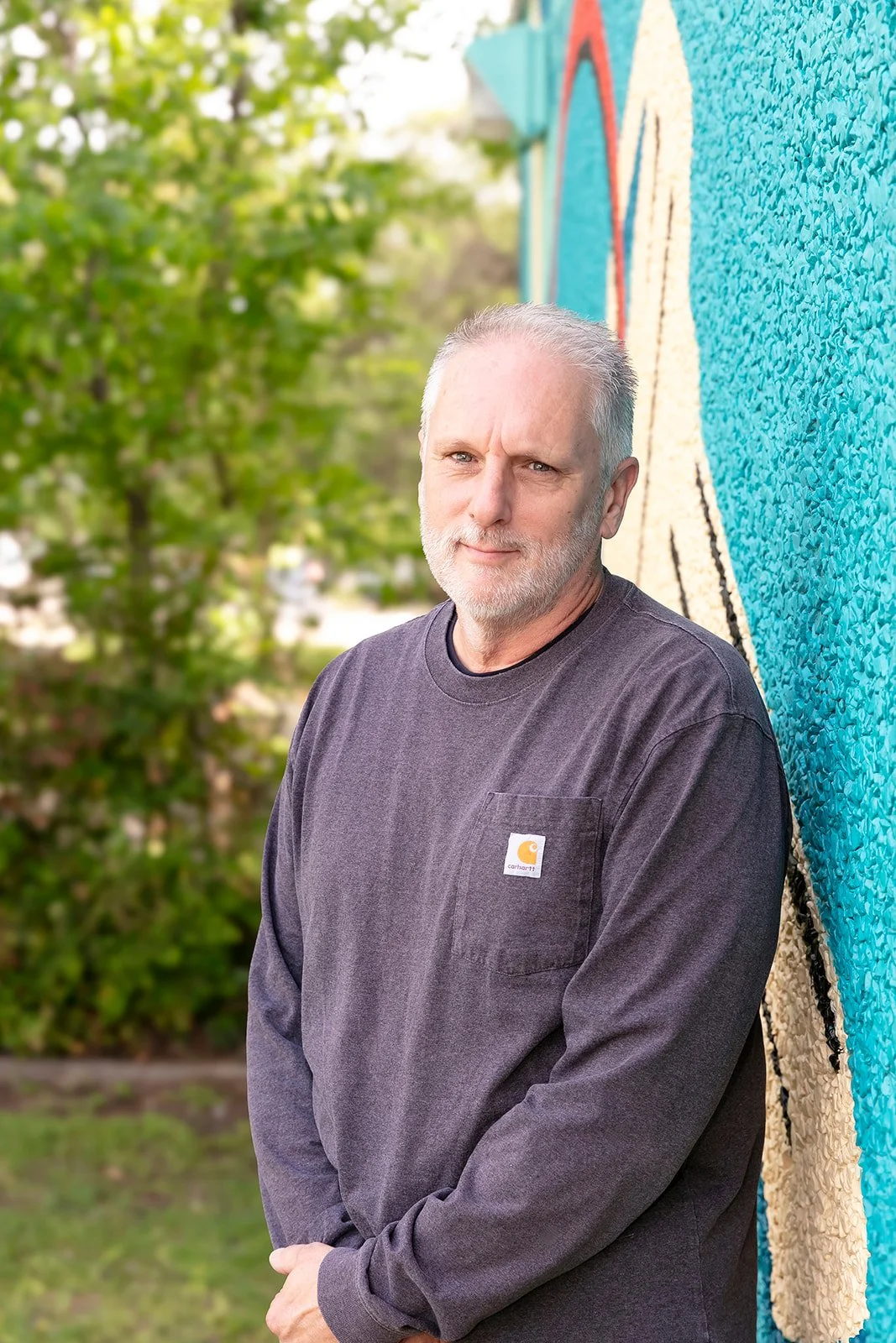 Close-up of a mature man with gray hair and beard, wearing a black shirt, standing in front of a mural.
