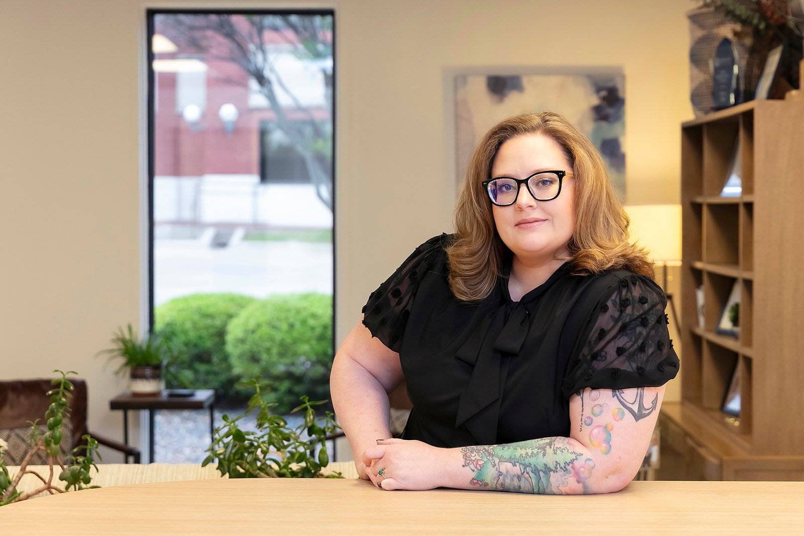 Ashley's headshot. A woman with shoulder-length red hair and glasses, wearing a black sleeveless top with a decorative bow on the shoulder, smiling in front of a brick wall.