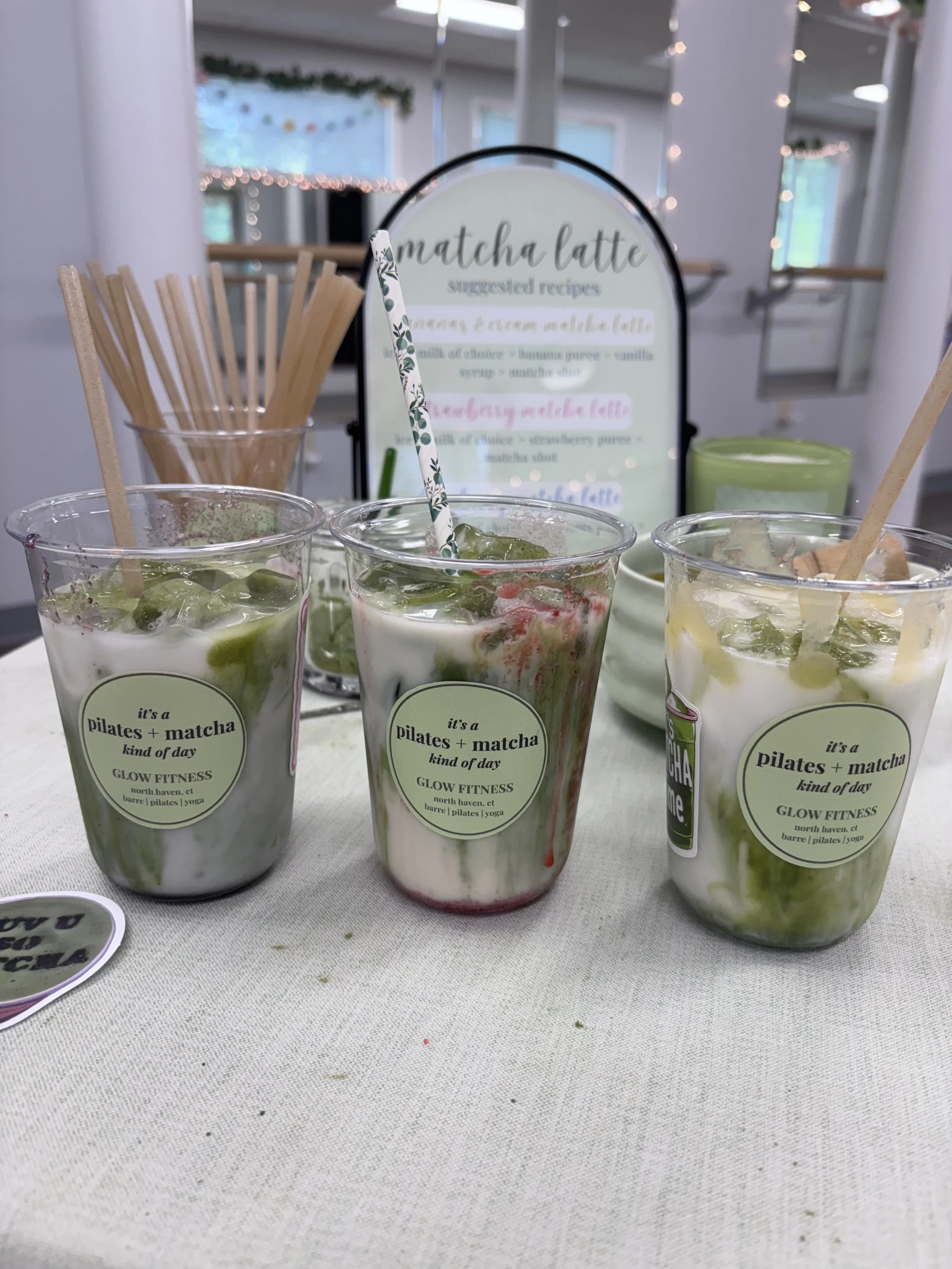 Three cups of matcha latte with whipped cream and green matcha powder, a container of wooden stir sticks, and a sign with suggested matcha latte recipes on a white table. The background shows a cozy indoor setting with string lights.