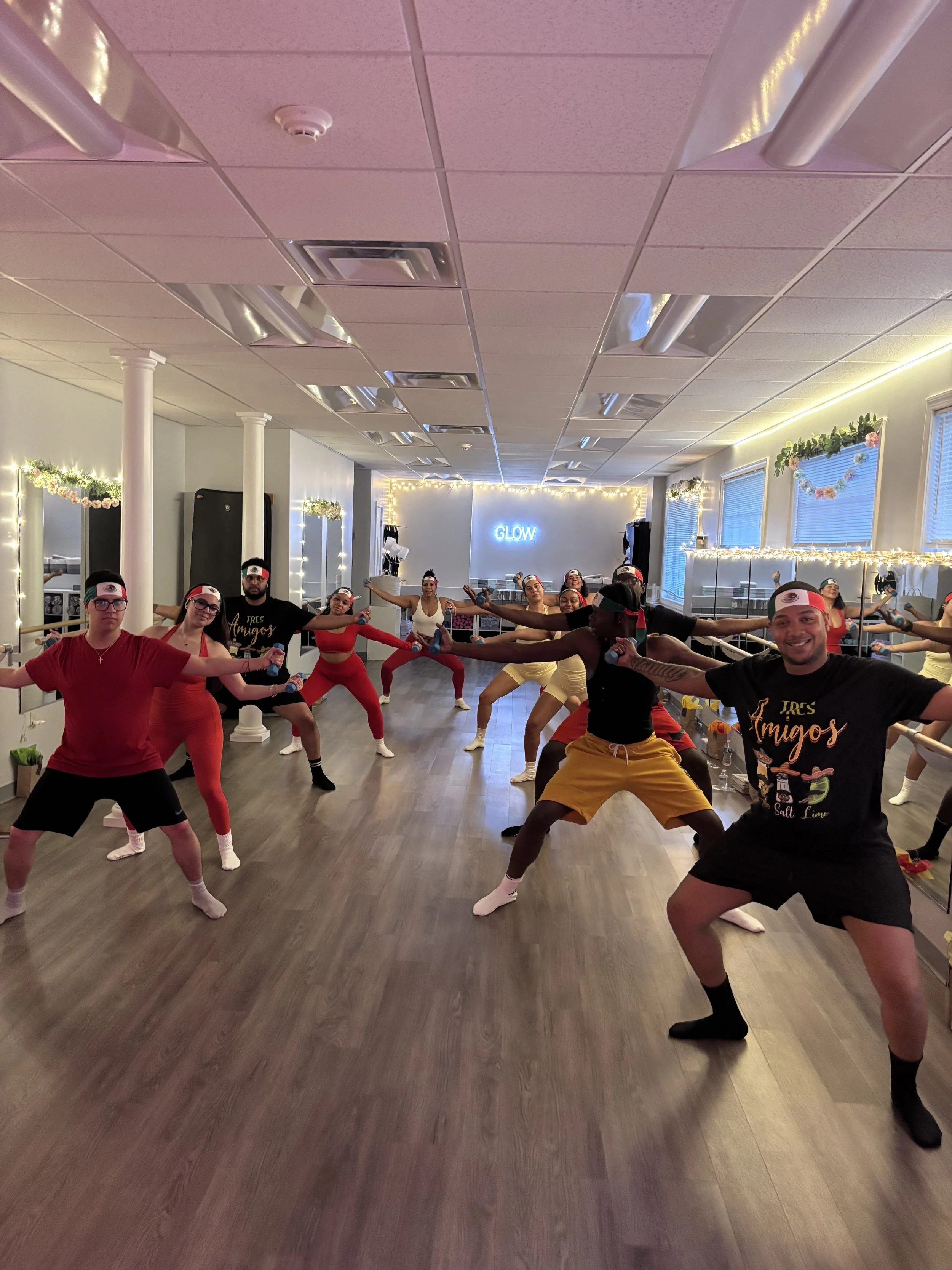 Group of diverse people in a dance studio doing a stretching or dance exercise, with a mirror, string lights, and a sign that says 'GLOW' in the background.