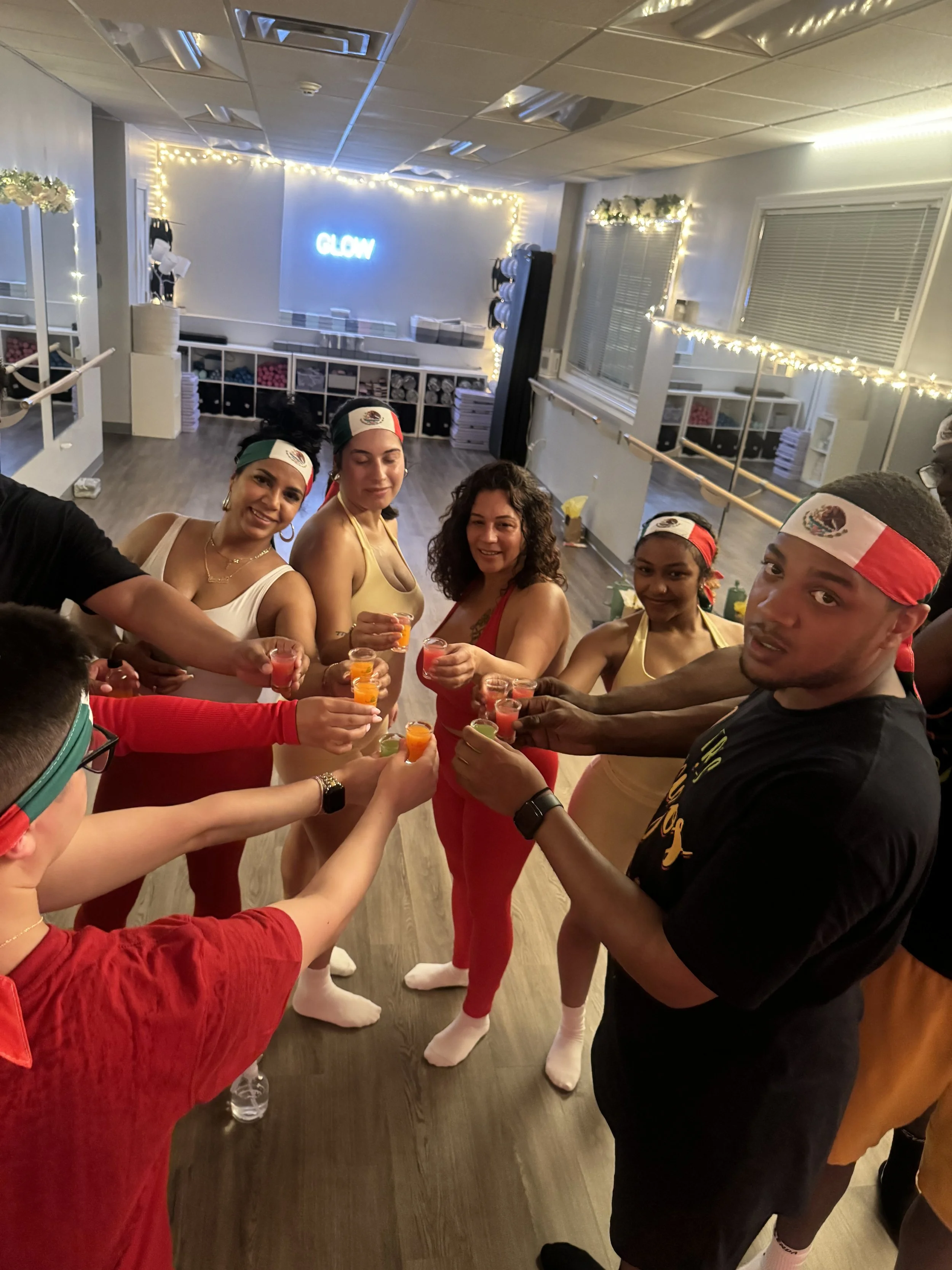 People celebrating and toasting with colorful shots in a decorated room with string lights, a mirrored wall, and a neon sign that says 'GLOW.'