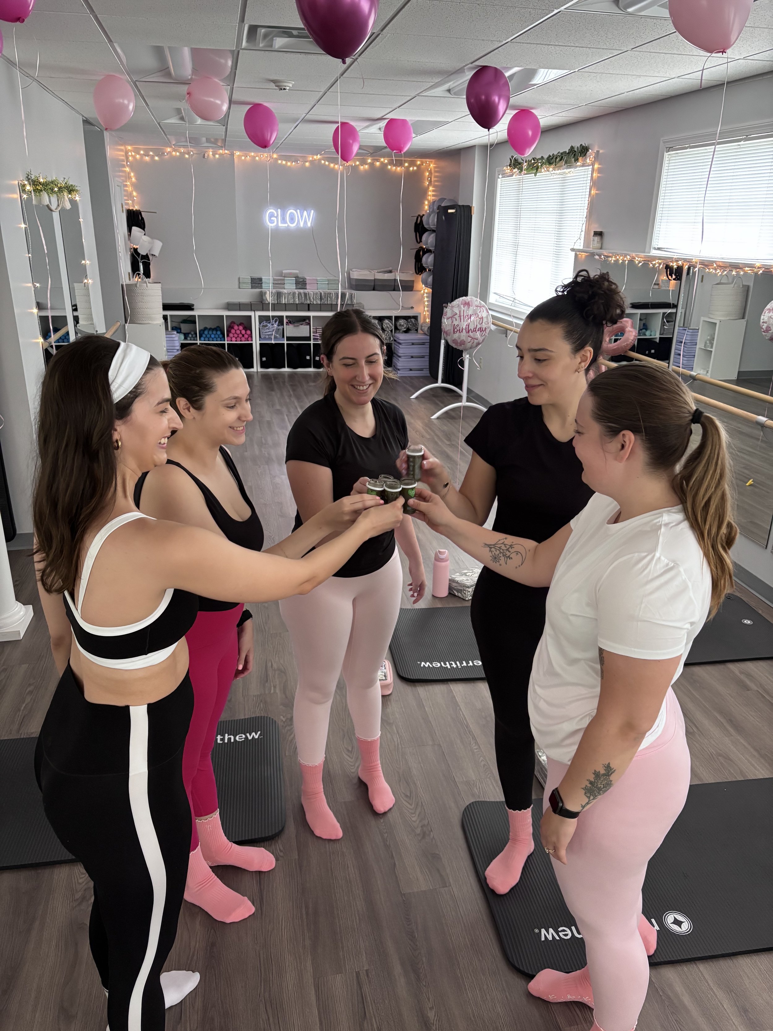 Five women at a birthday celebration, wearing athletic clothes and pink socks with cloud designs, toasting with small cups inside a decorated exercise studio with pink balloons, string lights, and a neon "GLOW" sign in the background.