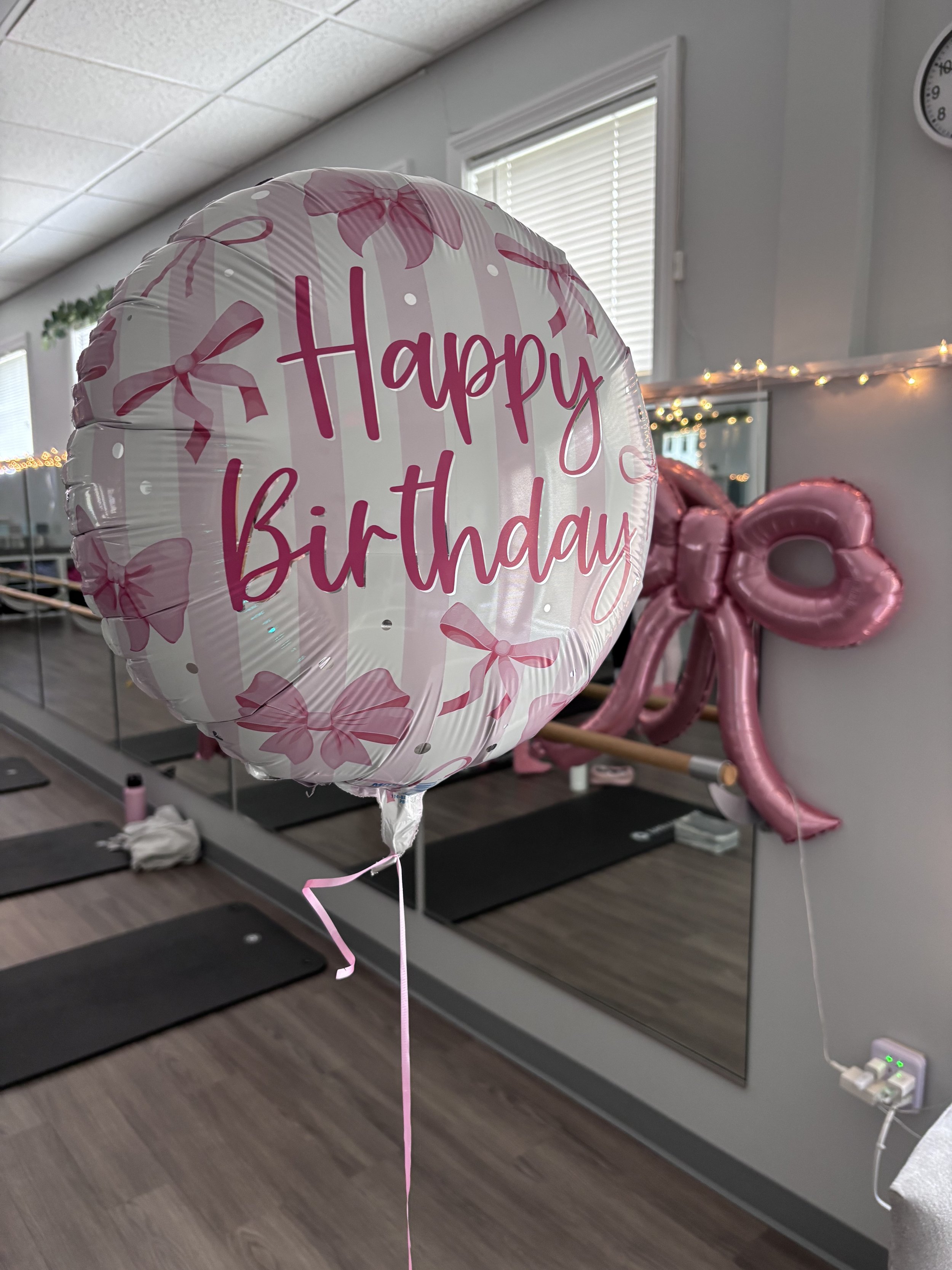 A pink and white balloon with the words 'Happy Birthday' written on it, decorated with pink bows, in a room with yoga mats, fairy lights, and windows.