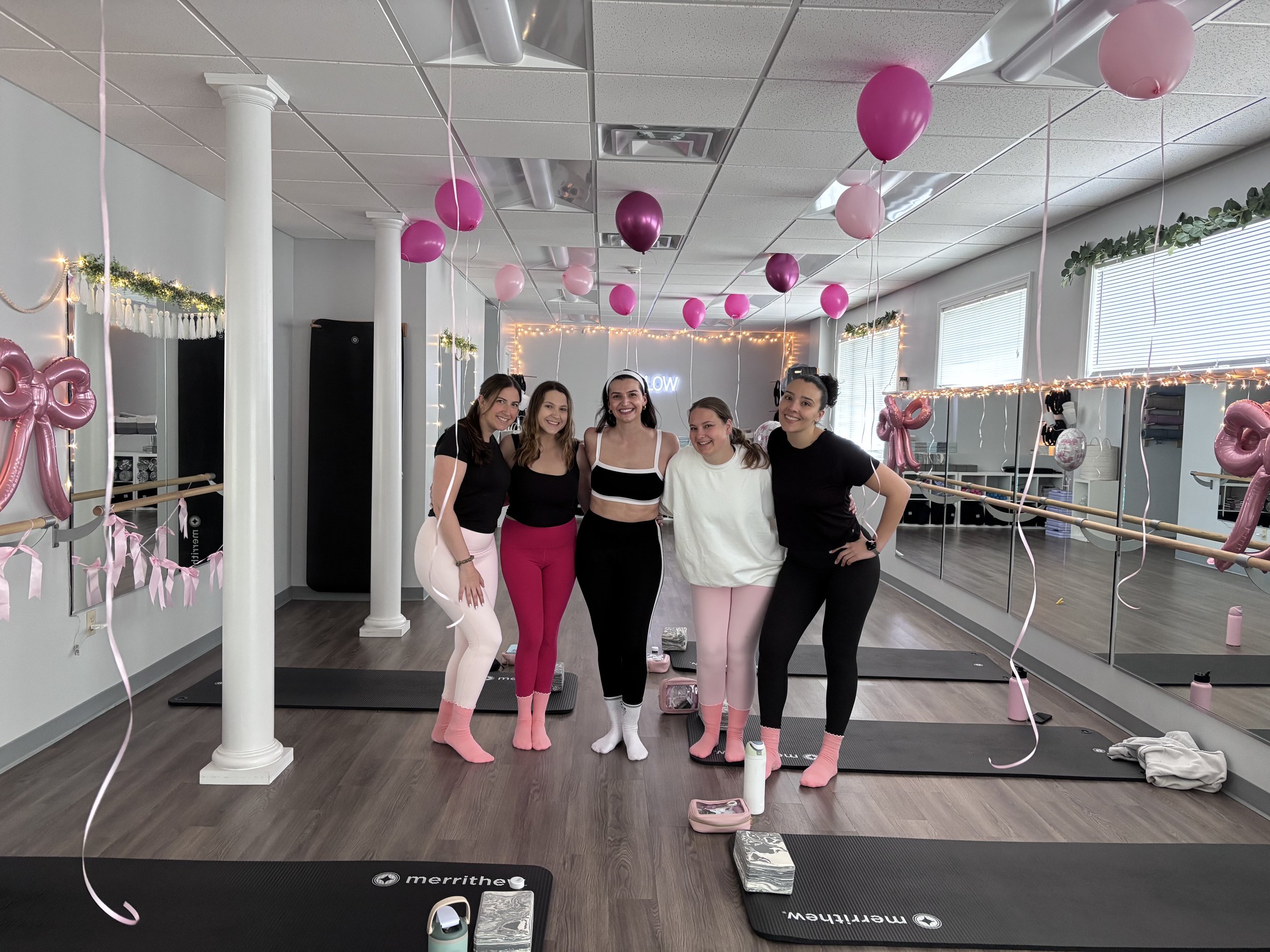 Group of five women dressed in casual athletic wear, standing together in a dance or fitness studio decorated with pink and purple balloons, fairy lights, and pink ribbon decorations, smiling at the camera.