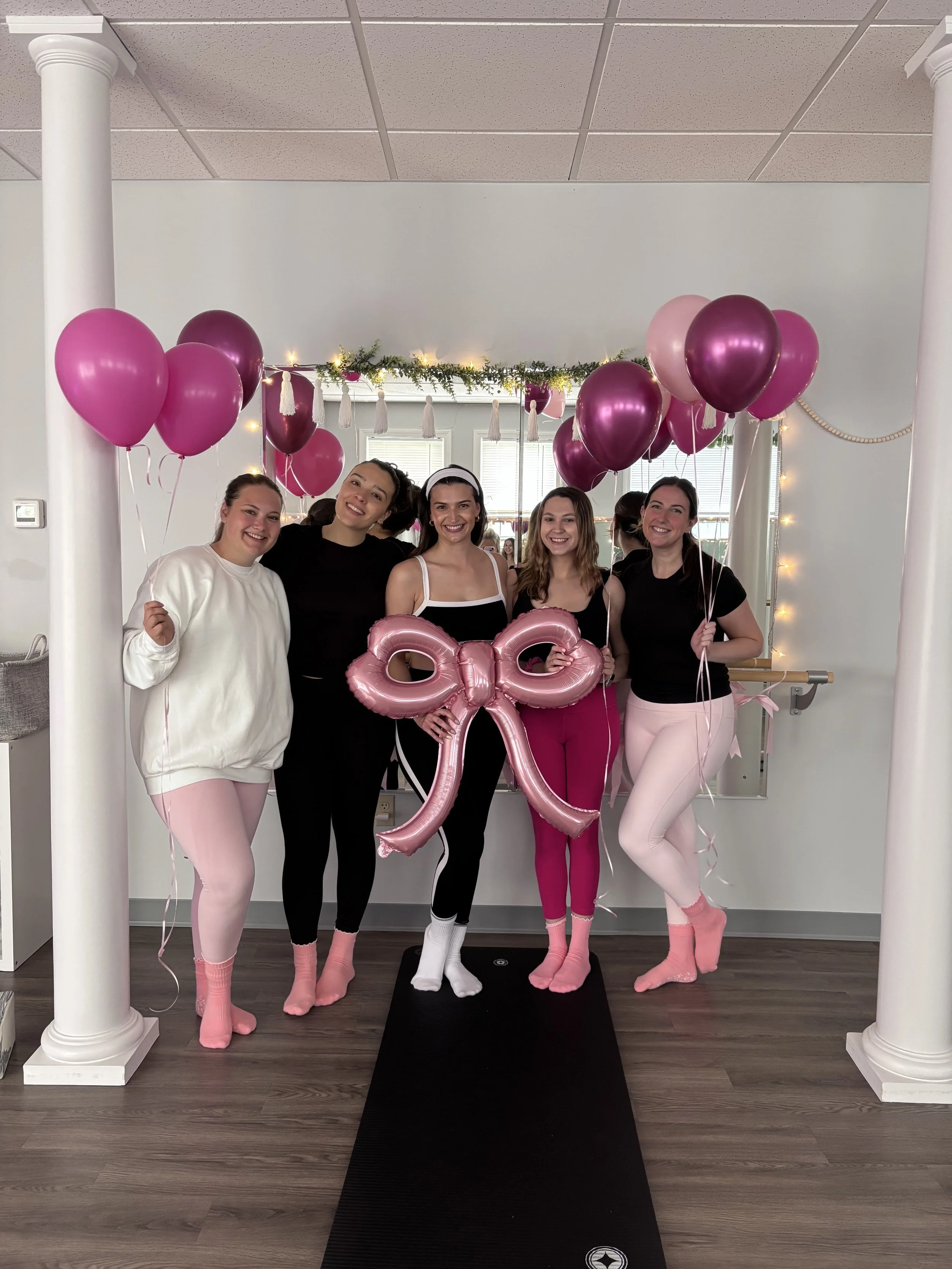 Group of five women standing on a black mat, holding pink and purple balloons, with a large pink bow balloon in the center, decorated with fairy lights, in a room with white walls and two white columns.