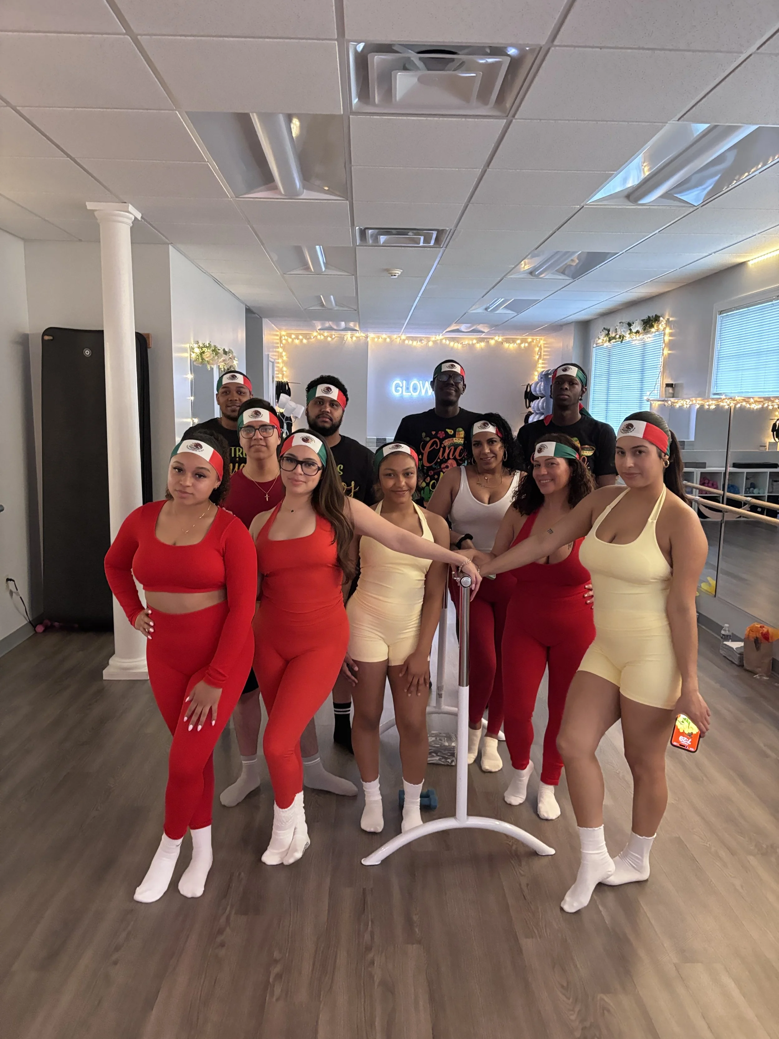 Group of ten people, mostly women, dressed in red, yellow, and black workout clothes with Mexican flag headbands, standing in a fitness or dance studio with wood floors and fairy lights. They are holding hands in front of a white disco ball stand.