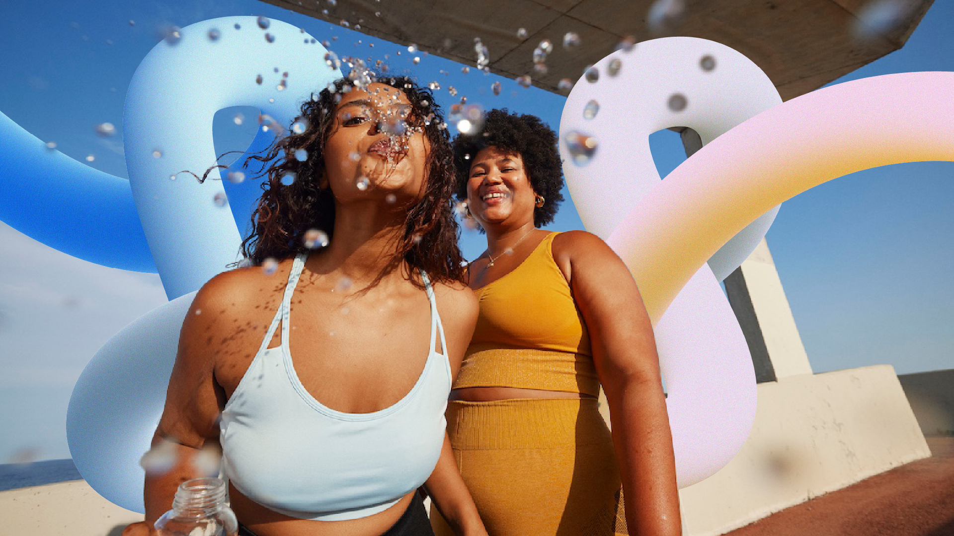 Two women enjoying a day at the beach with large colorful balloons in the background, one woman blowing water from a bottle, both smiling and having fun.