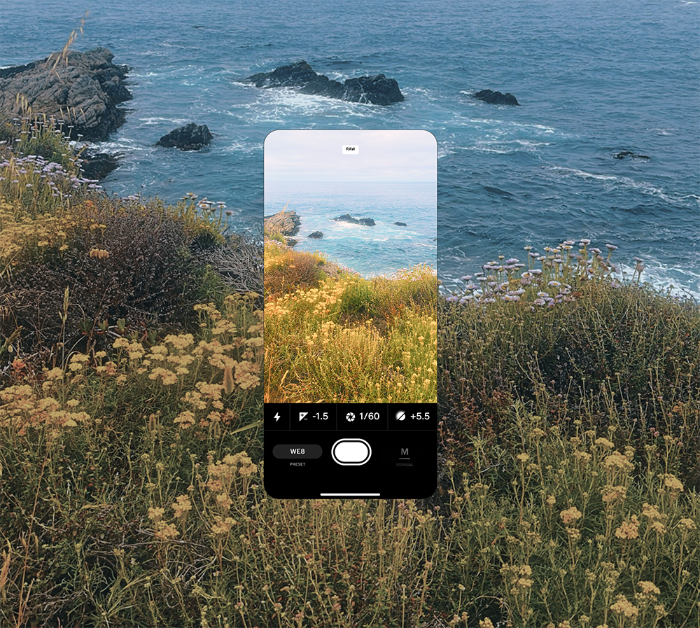 The image features a coastal scene with a rugged shoreline and the ocean in the background. A smartphone screen is superimposed over the scene, showing the camera settings and a live view of the coastline and wildflowers in the foreground. The phone's view highlights a portion of the landscape with plants and rocks near the water.