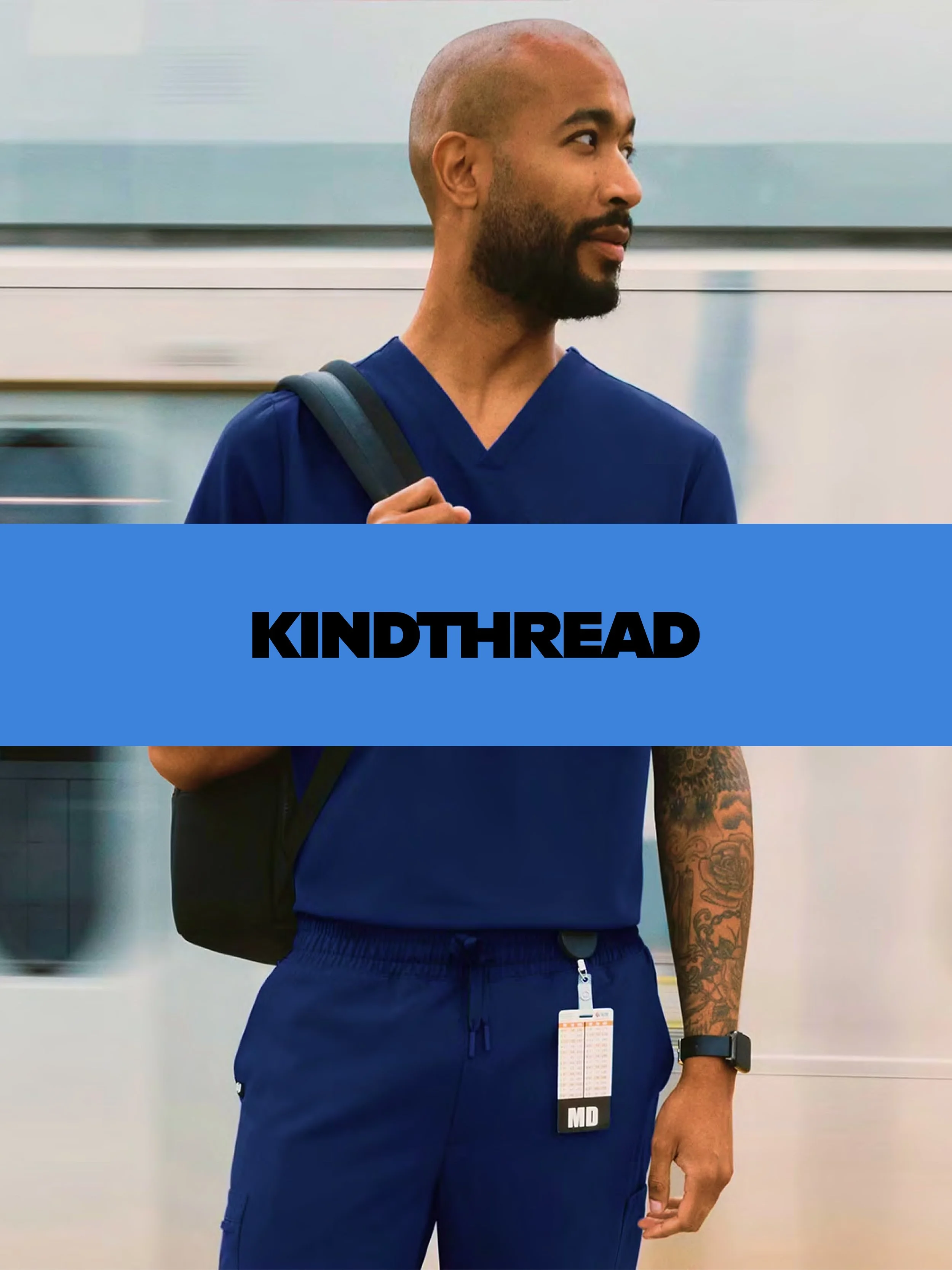 A man in blue scrubs standing in front of a moving train with a backpack, a watch, and an ID badge, holding a strap over his shoulder, on a train platform.
