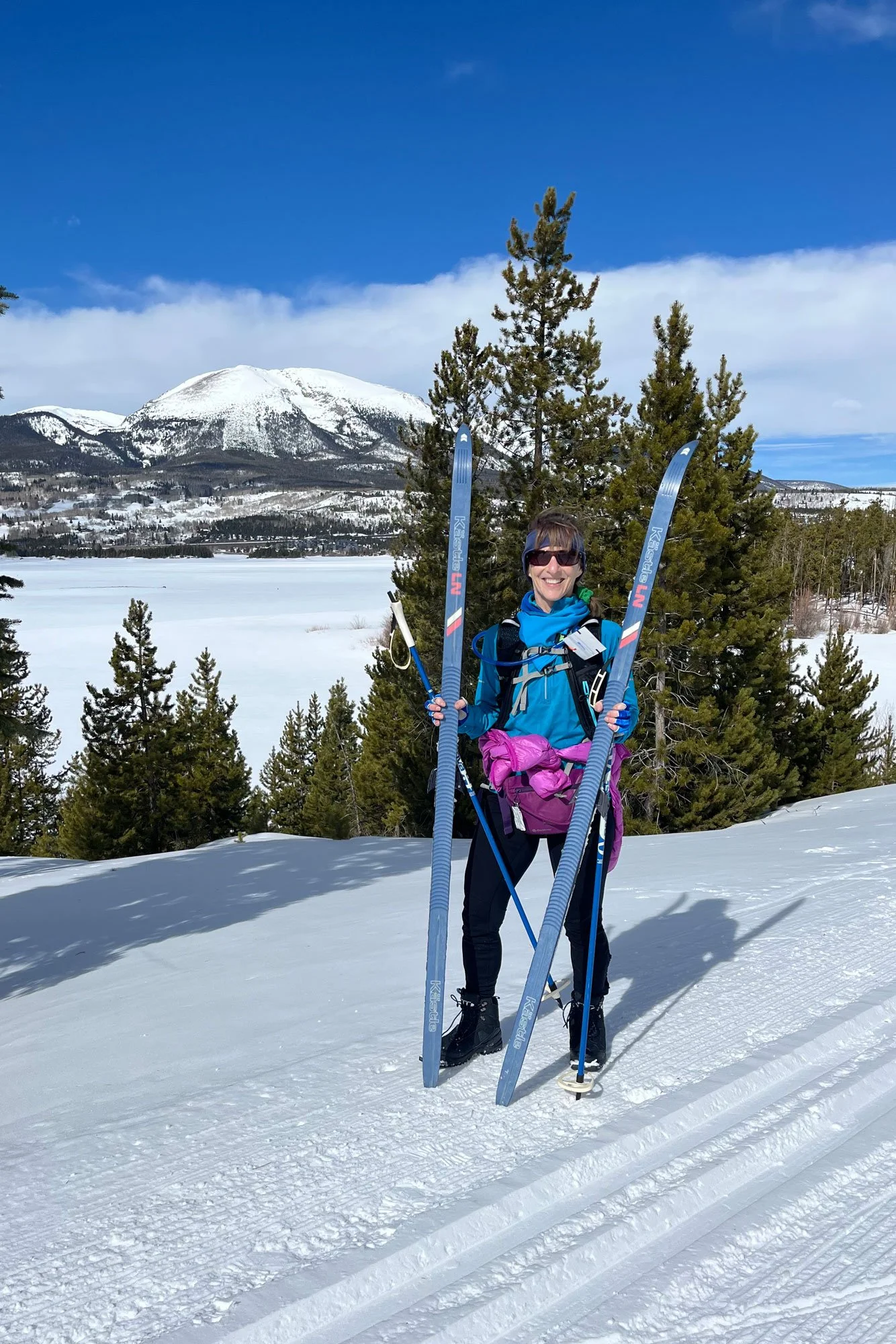 A woman smiling in a snowy landscape holding cross-country skis and ski poles, with a mountain and pine trees in the background under a blue sky.