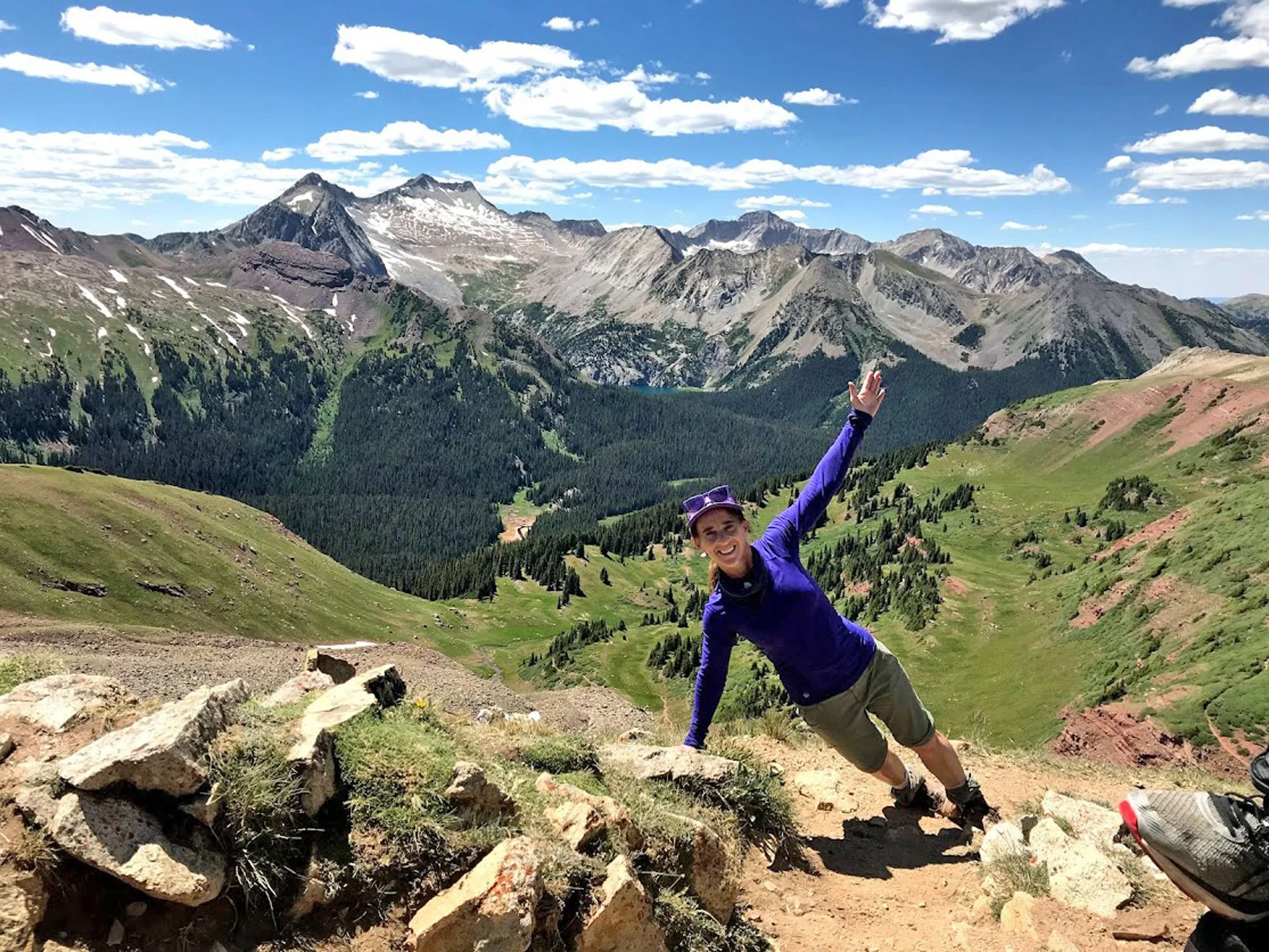 A smiling woman in hiking gear does a side plank on a mountain trail, with a scenic backdrop of green valleys, rugged mountain peaks, and a partly cloudy sky.