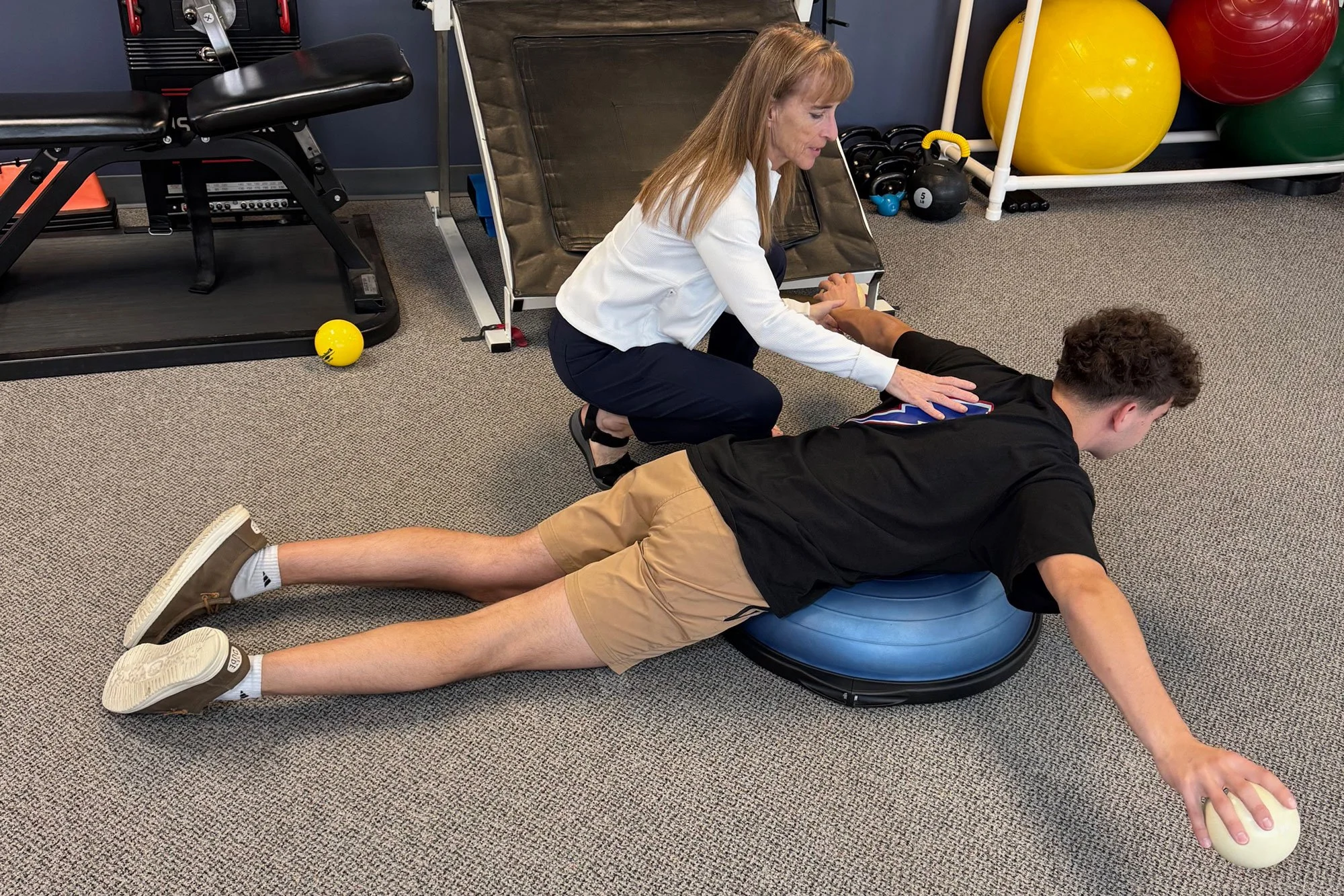 A physical therapy session in a gym, where a woman is assisting a young man with an exercise on a blue balance disc. The woman is kneeling beside the man, guiding his arm extension, while the young man is lying face down with his arm extended forward, holding a small weight. The background includes gym equipment, exercise balls, and kettlebells.
