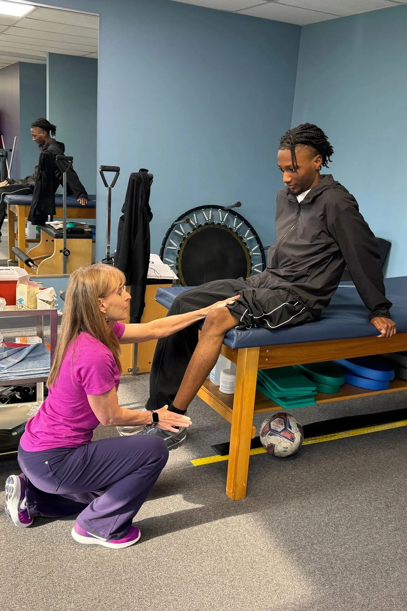 A physical therapysession where a woman in a pink shirt kneels on the floor supporting a man's leg on a padded table. The man, dressed in black athletic clothing, sits on the table with his leg extended. A healthcare professional or trainer assists with the exercise. Medical and exercise equipment is visible in the background.