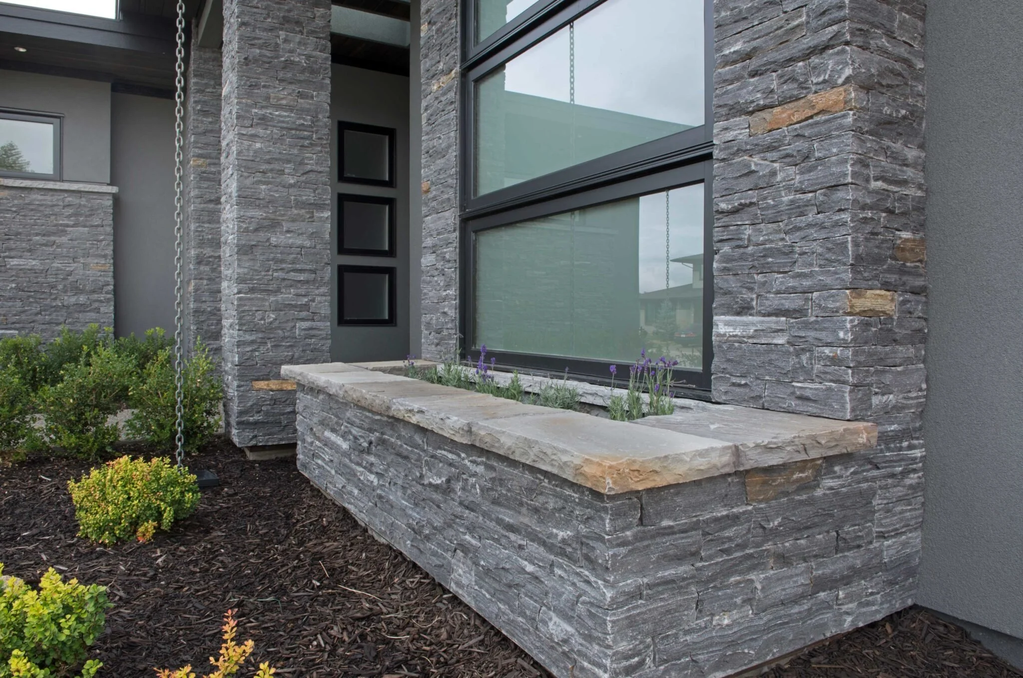 Close-up of a modern house exterior with gray stacked stone siding, large windows, a stone window box with lavender flowers, and landscaped garden with mulch and green bushes.