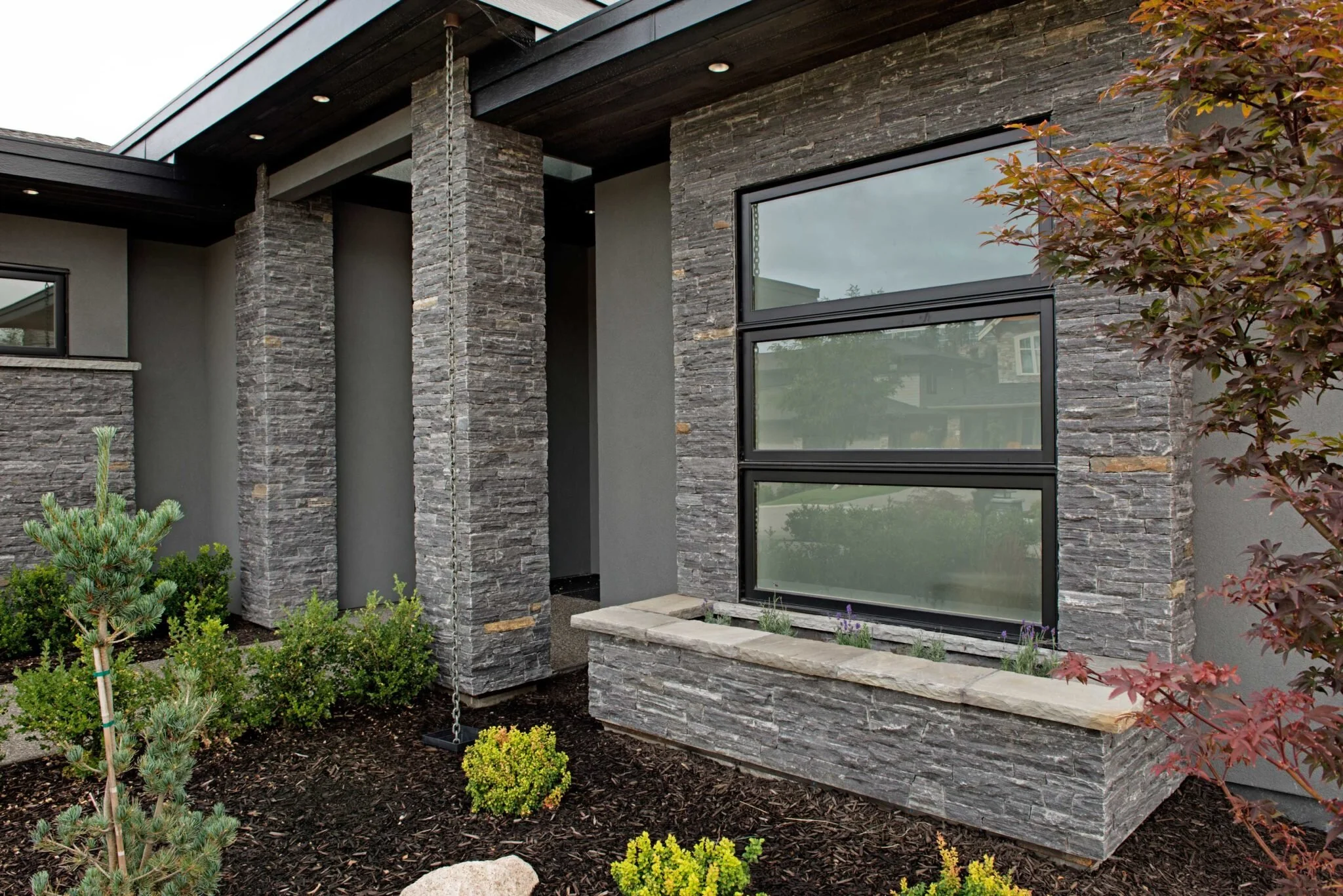 Modern house exterior with stone and stucco walls, large rectangular window, landscaped garden with small bushes and mulch.