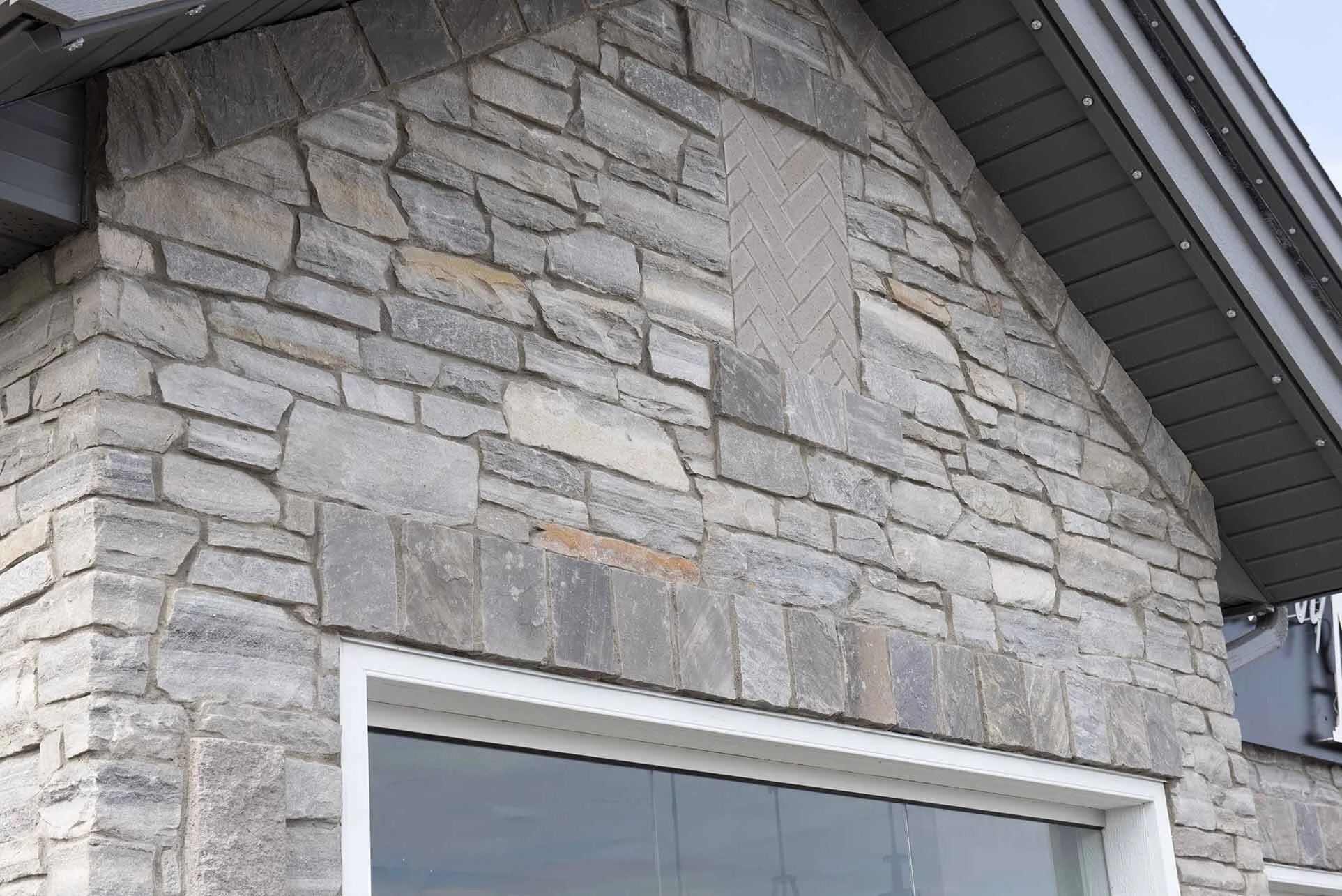 Close-up of a stone exterior wall of a building with a window, showing gray and beige stones, some arranged in a herringbone pattern, under a dark metal roof.